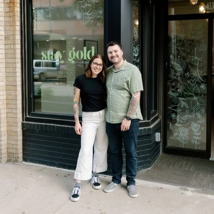 A woman and man standing together outside a storefront with large windows and floral illustrations. The woman wears a black shirt and white wide-leg pants, and the man wears a green patterned shirt and dark jeans. They are both smiling.