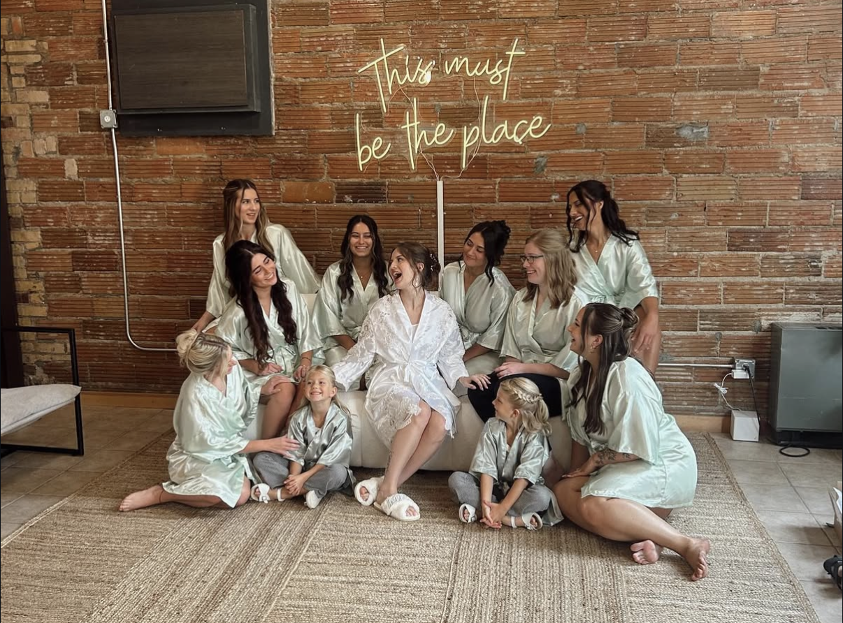 A group of women and girls in satin robes gathered in a cozy, rustic room with an exposed brick wall. They are smiling and laughing, with some sitting on the floor and others on a white couch. There is a neon sign on the wall that reads, "This must be the place." The atmosphere appears joyful and celebratory.