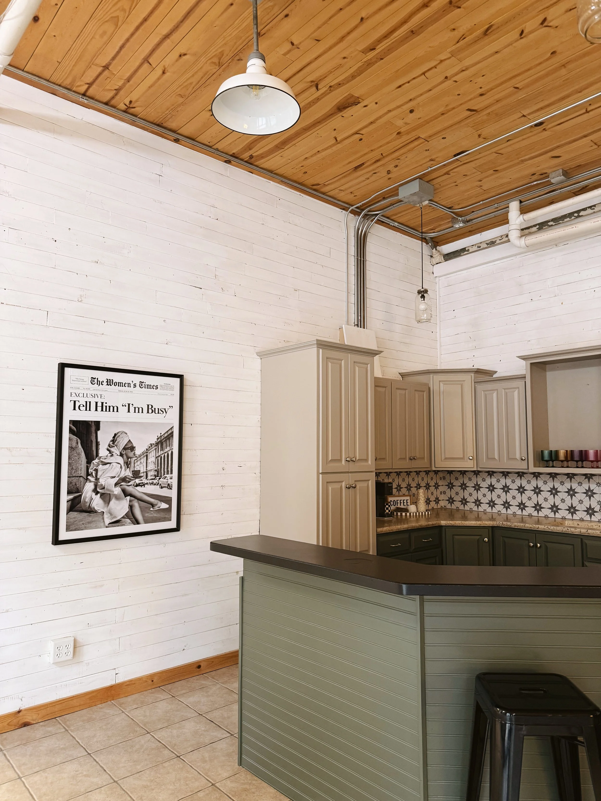 A corner of a kitchen with beige cabinets and a black countertop, a black stool, a black and white framed photo on the white wooden wall, a potted plant, and colorful cups on a shelf. The ceiling is wooden with exposed pipes and a light fixture.
