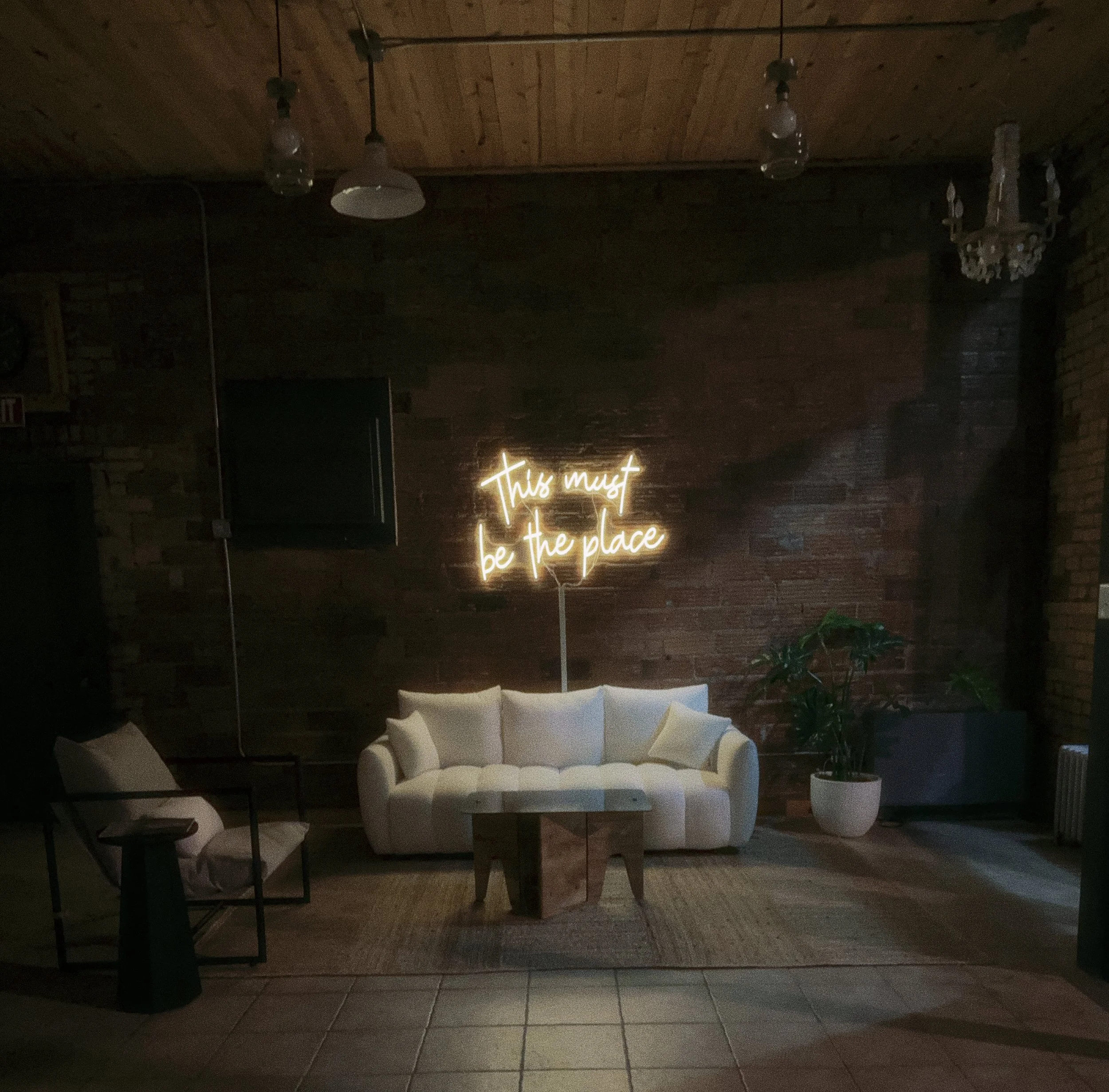 Interior of a cozy lounge area with a white sofa, a small wooden coffee table, a chair with a side table, potted plant, and a neon sign that reads 'This must be the place' on a brick wall.