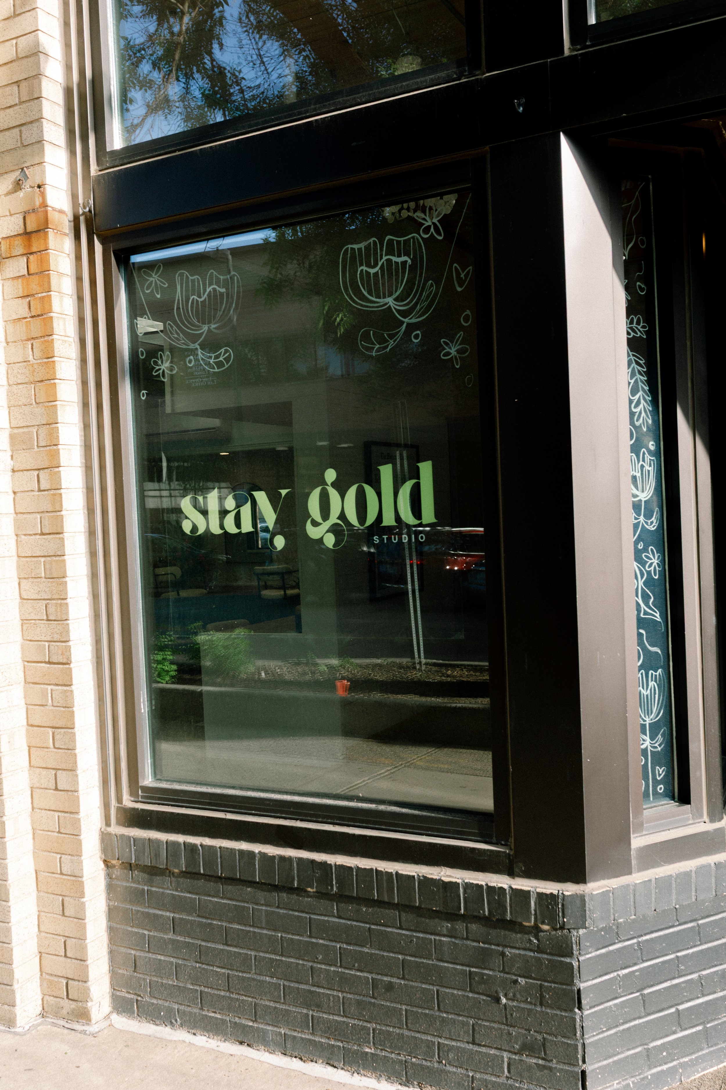 Storefront window with green text that says 'stay gold' and the word 'studio' beneath it, surrounded by white line drawings of flowers and leaves.