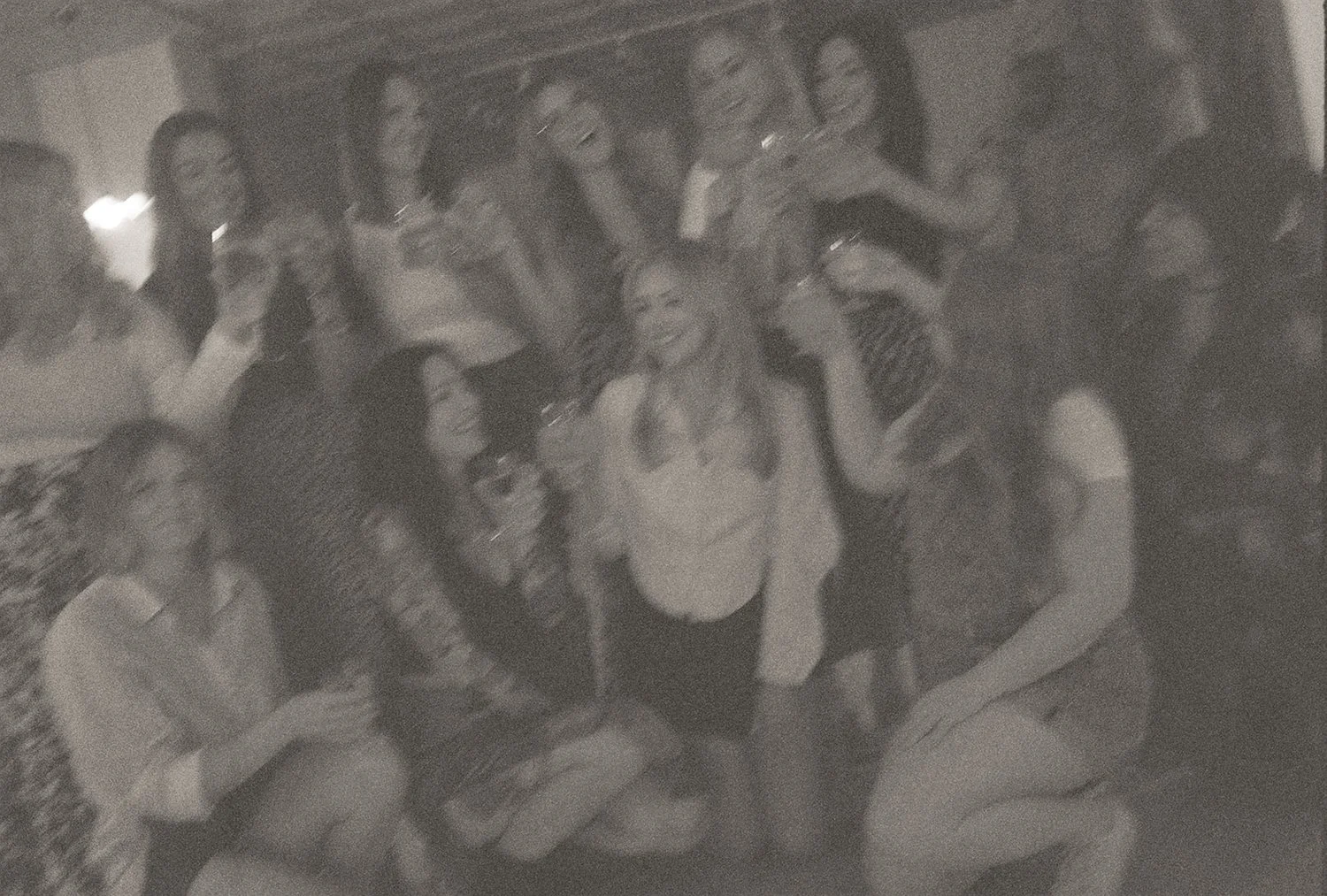 Group of women celebrating with drinks, smiling and laughing, in a blurred grayscale photograph.