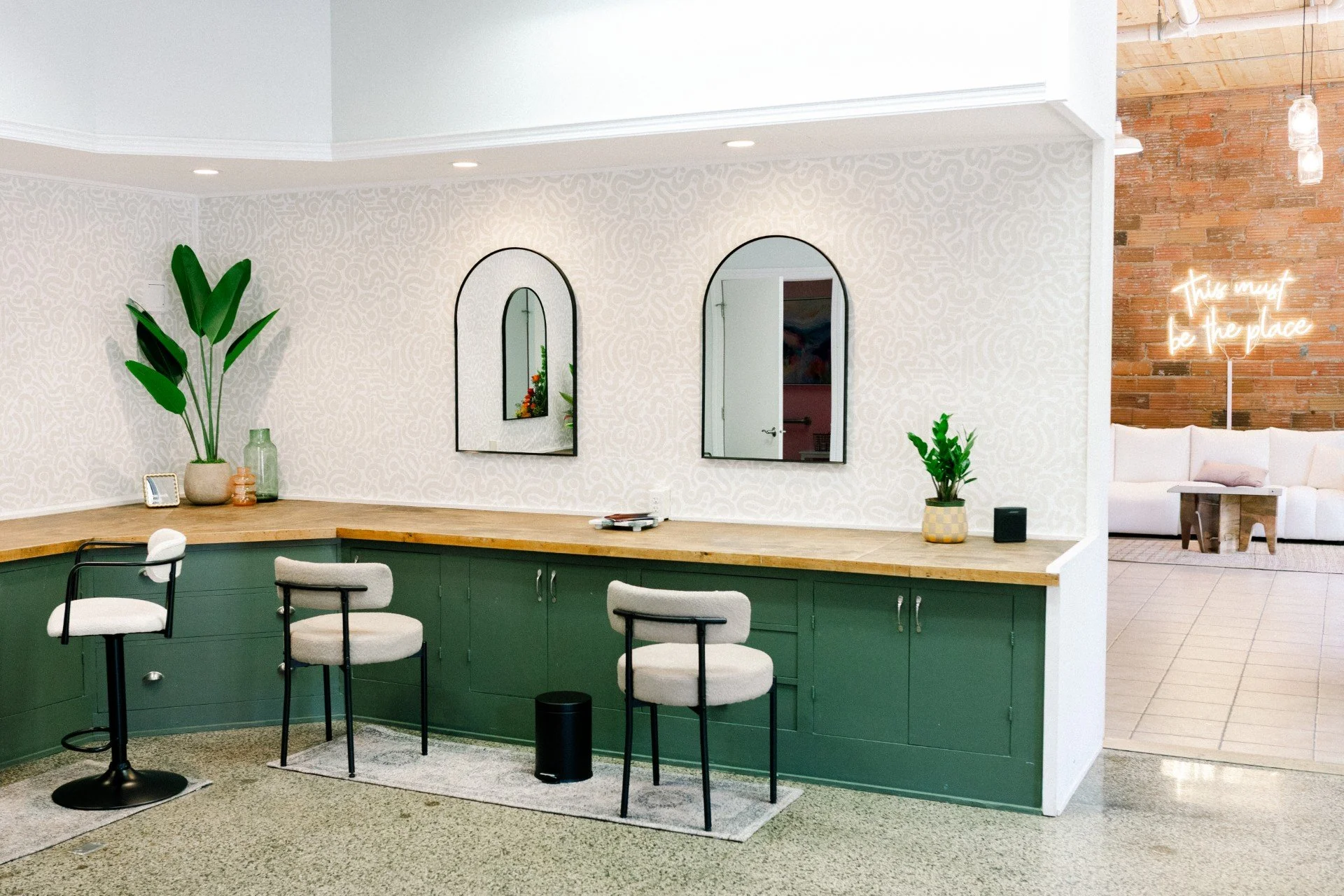 Modern interior space with a curved green counter, white upholstered chairs, decorative plants, two arched mirrors on the wall, and a neon sign reading "This must be the place" on a brick wall.