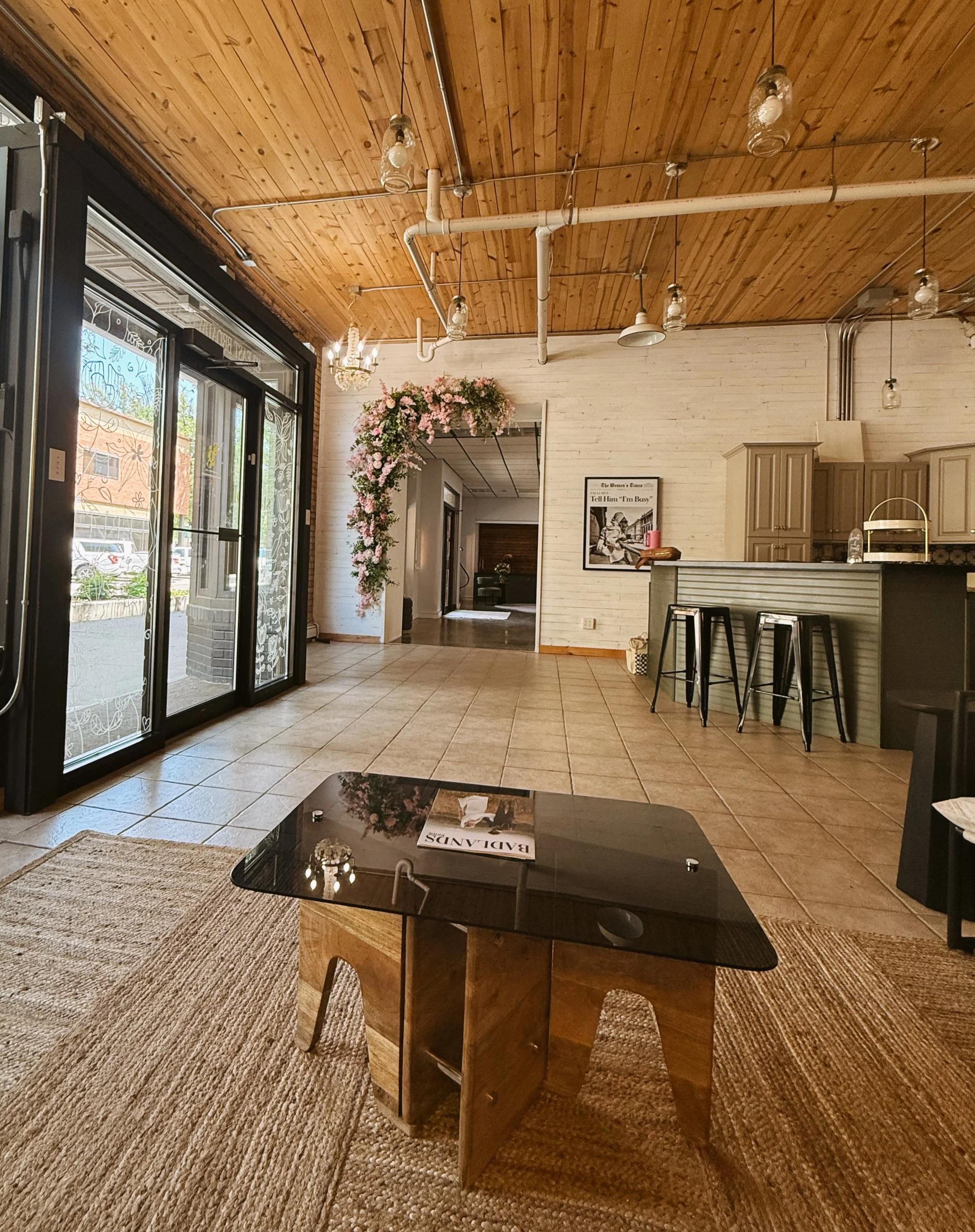 Interior of a cafe or lounge with a wooden ceiling, tiled floor, black glass-top coffee table on a woven rug, bar counter with three black stools, floral arrangement, and framed art on white brick wall.