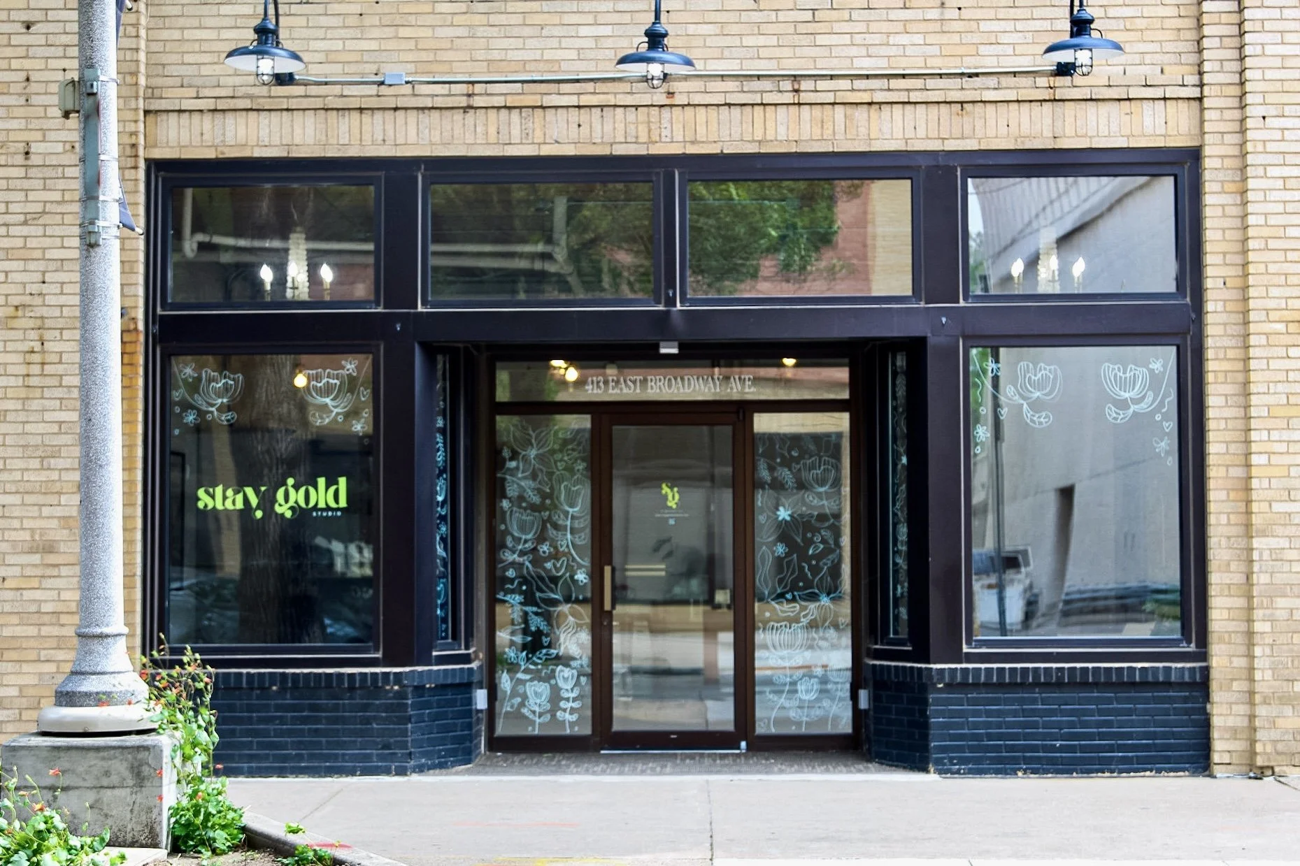 Storefront with black framing, large windows with floral decorations, neon sign that reads 'stay gold,' and an address sign that says '413 East Broadway Ave.'