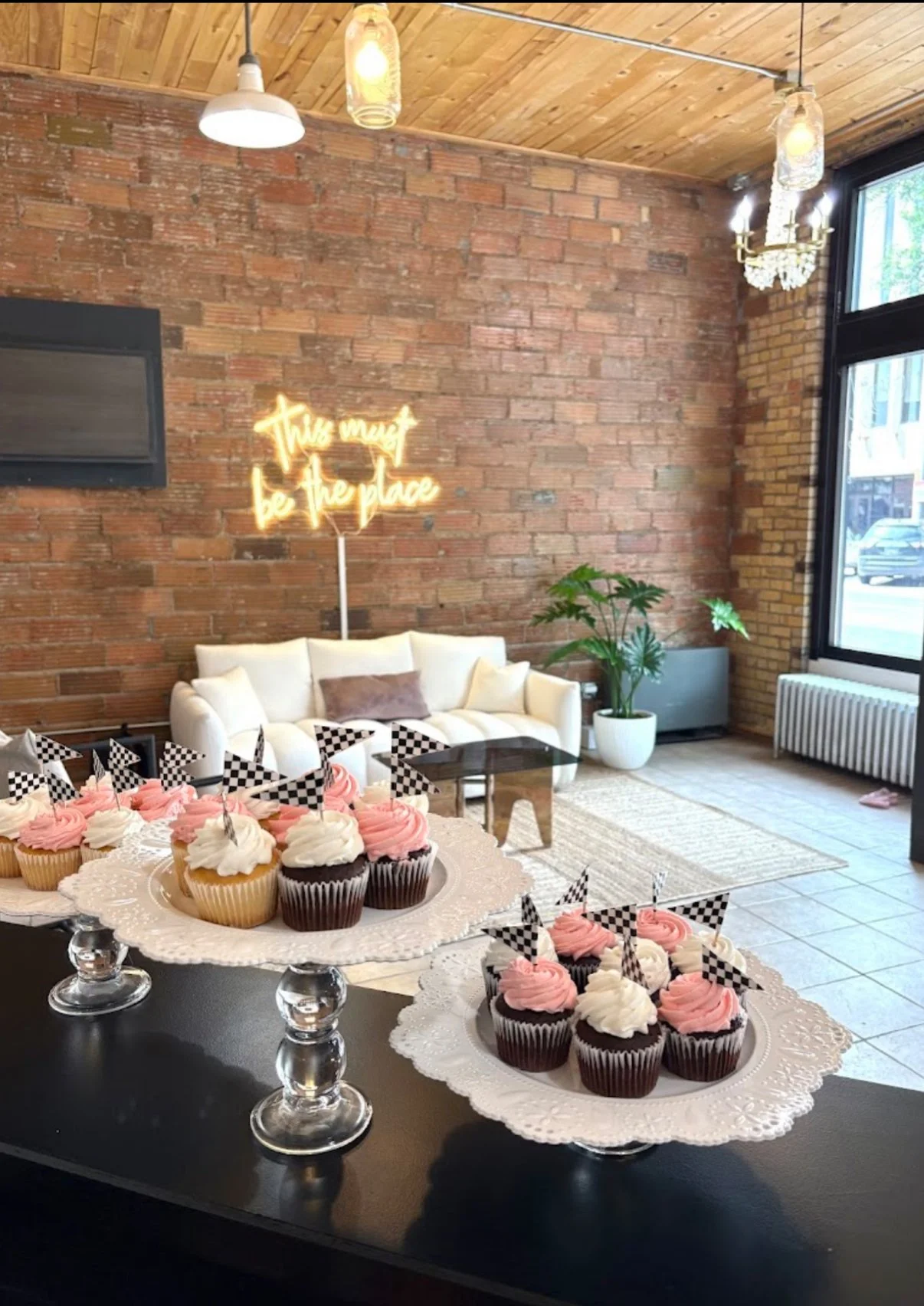 Cupcakes with pink and white frosting and checkered flags on a table, with a cozy living room in the background featuring a white couch, brick walls, large windows, and a neon sign that says 'This must be the place'.