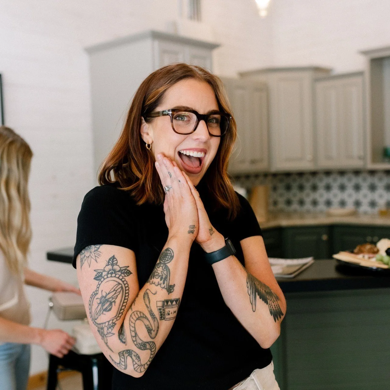 A woman with tattoos, glasses, and shoulder-length brown hair, smiling and holding her hands to her face in an indoor setting with a kitchen in the background.