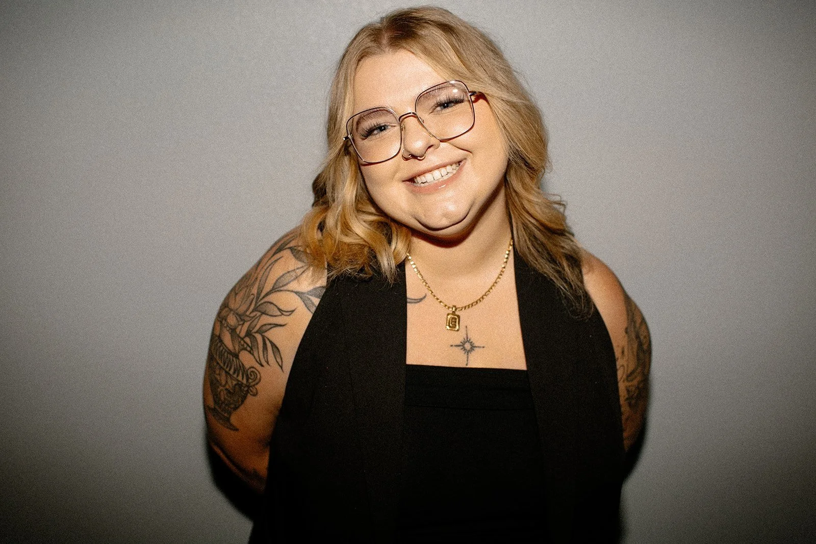 Smiling person with blonde hair, wearing glasses and a black top, showing tattoos on their arms, posed against a plain background.