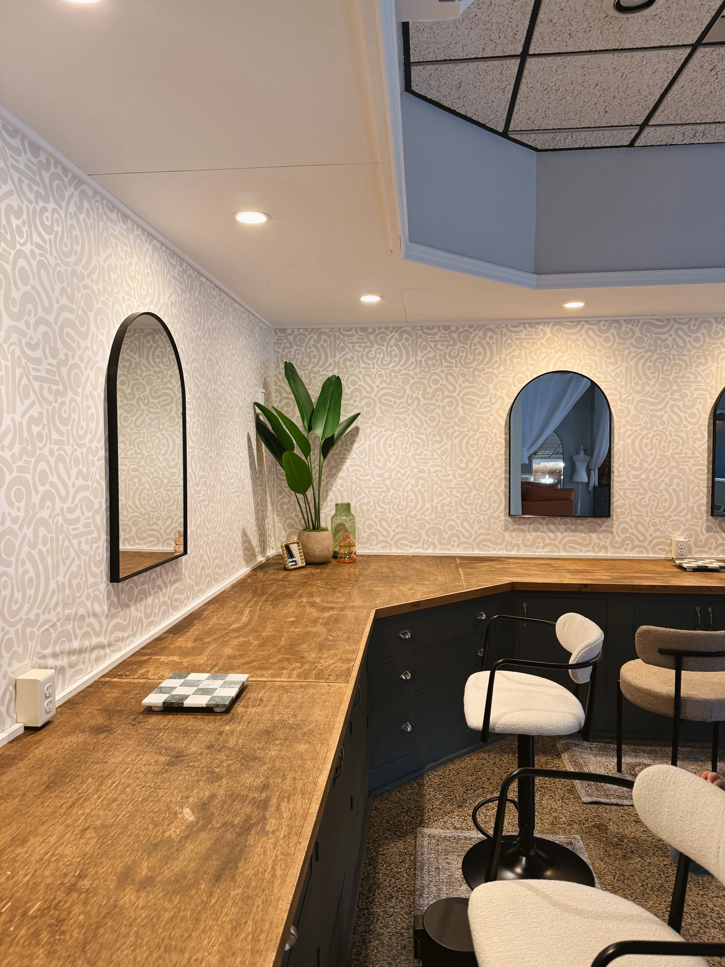 Corner of a modern room with a wooden countertop, two round white chairs with armrests, plants, and mirrors on the wall.