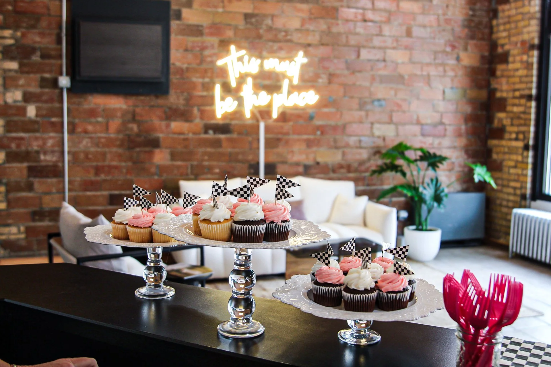 Cupcakes with pink and white frosting on silver cake displays in a cozy indoor setting with a brick wall, green plants, and a neon sign that says 'this moment be the place'.