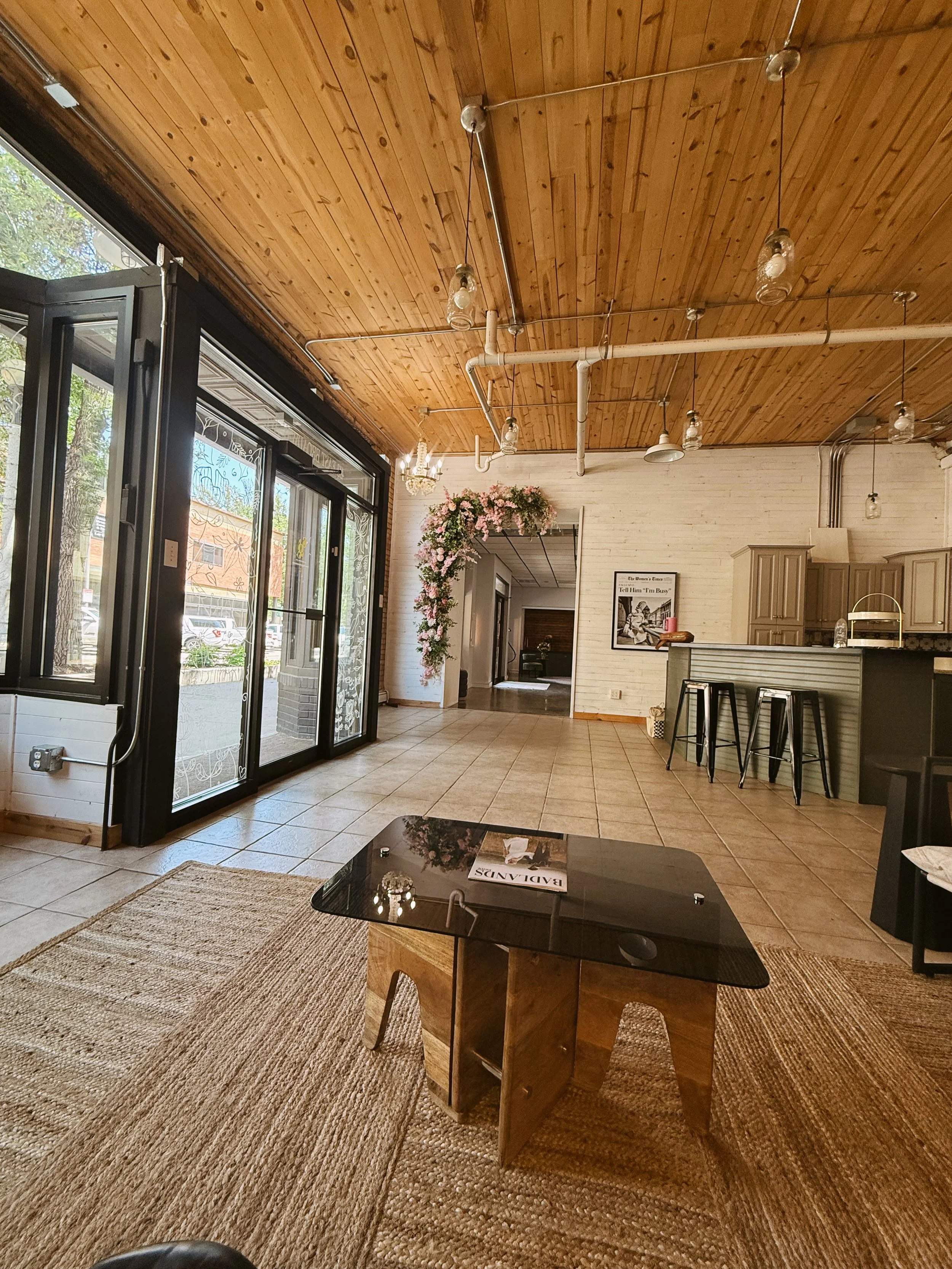 Interior of a cozy studio with a wooden ceiling, large front windows, a small black table on a woven rug, a welcoming floral archway, and a bar area with three black stools.
