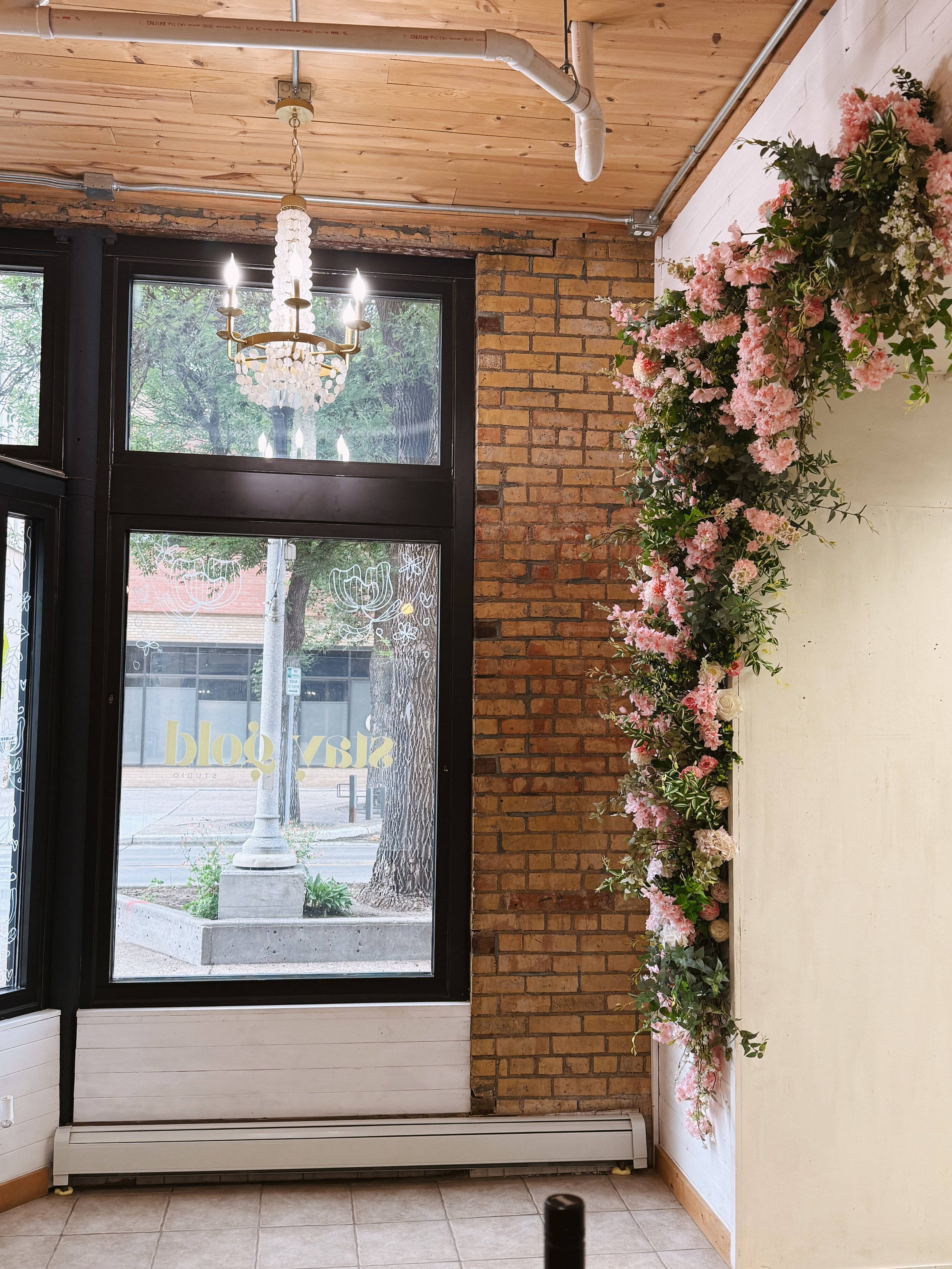 Interior of a room with a large window, a chandelier, and pink flowers arranged along the wall.