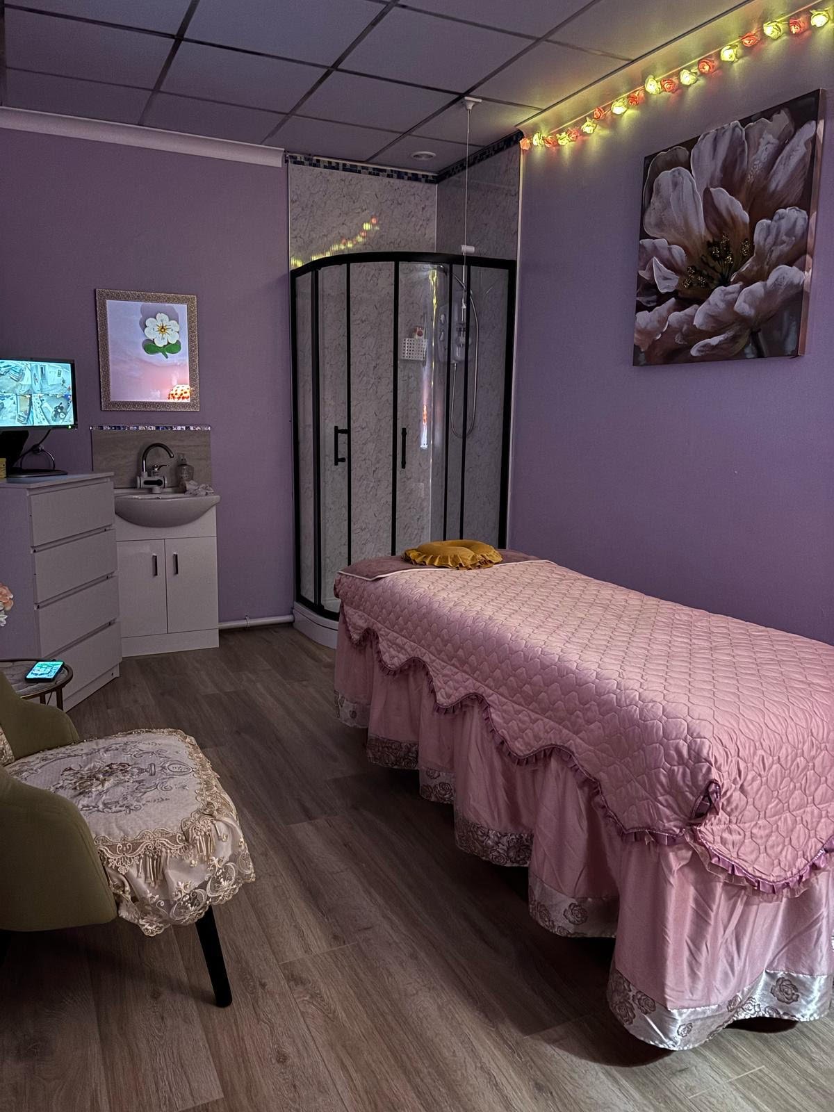 A cozy room with a massage table covered in a pink quilted blanket. The walls are light purple, adorned with a large floral painting. There is a white sink cabinet and a glass shower enclosure in the corner. Soft lighting from string lights enhances