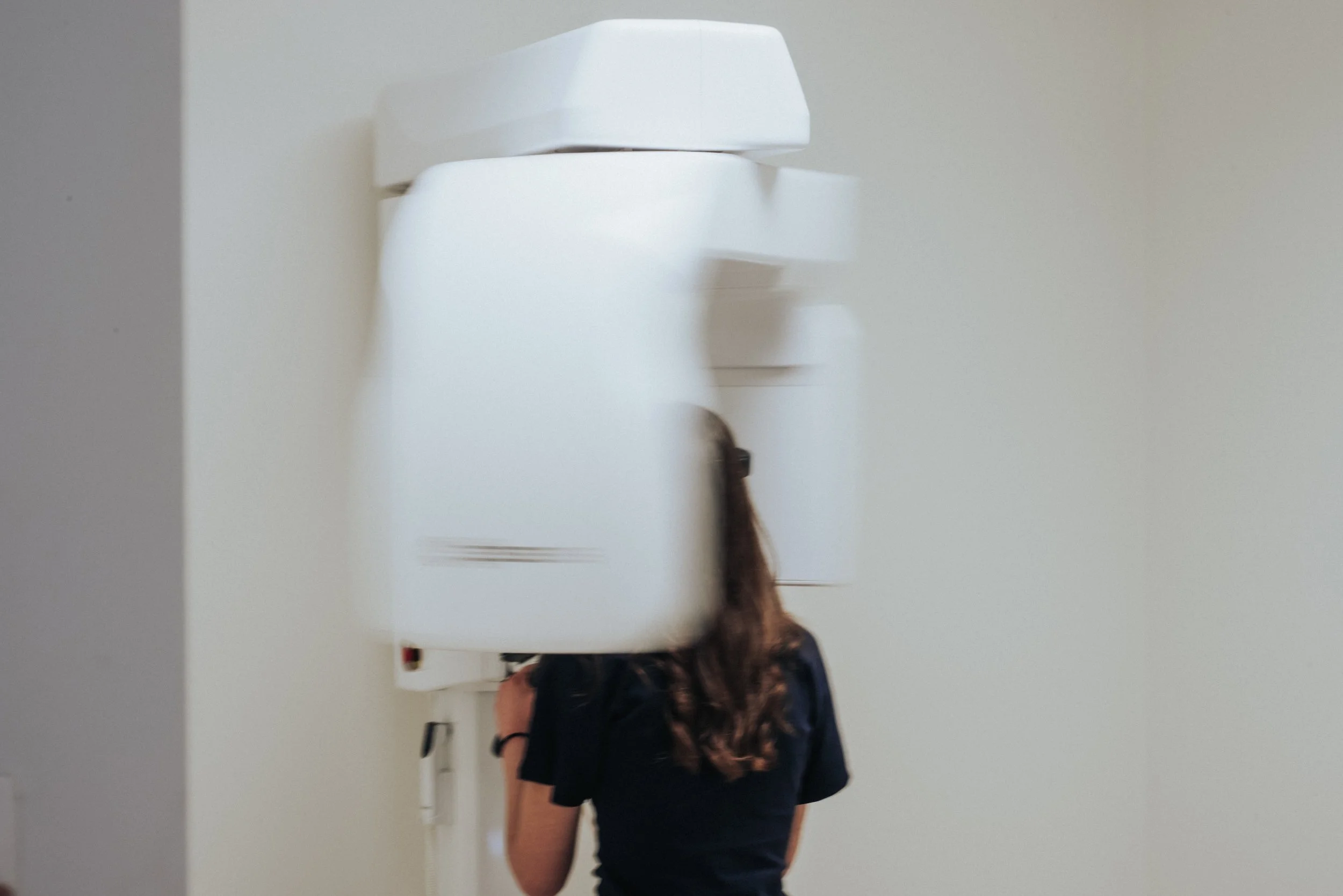 A woman with long brown hair wearing a black shirt and black shorts, seen from the back, standing in front of a white wall and a mounted white object that appears to be an air conditioning unit or sanitizer dispenser.