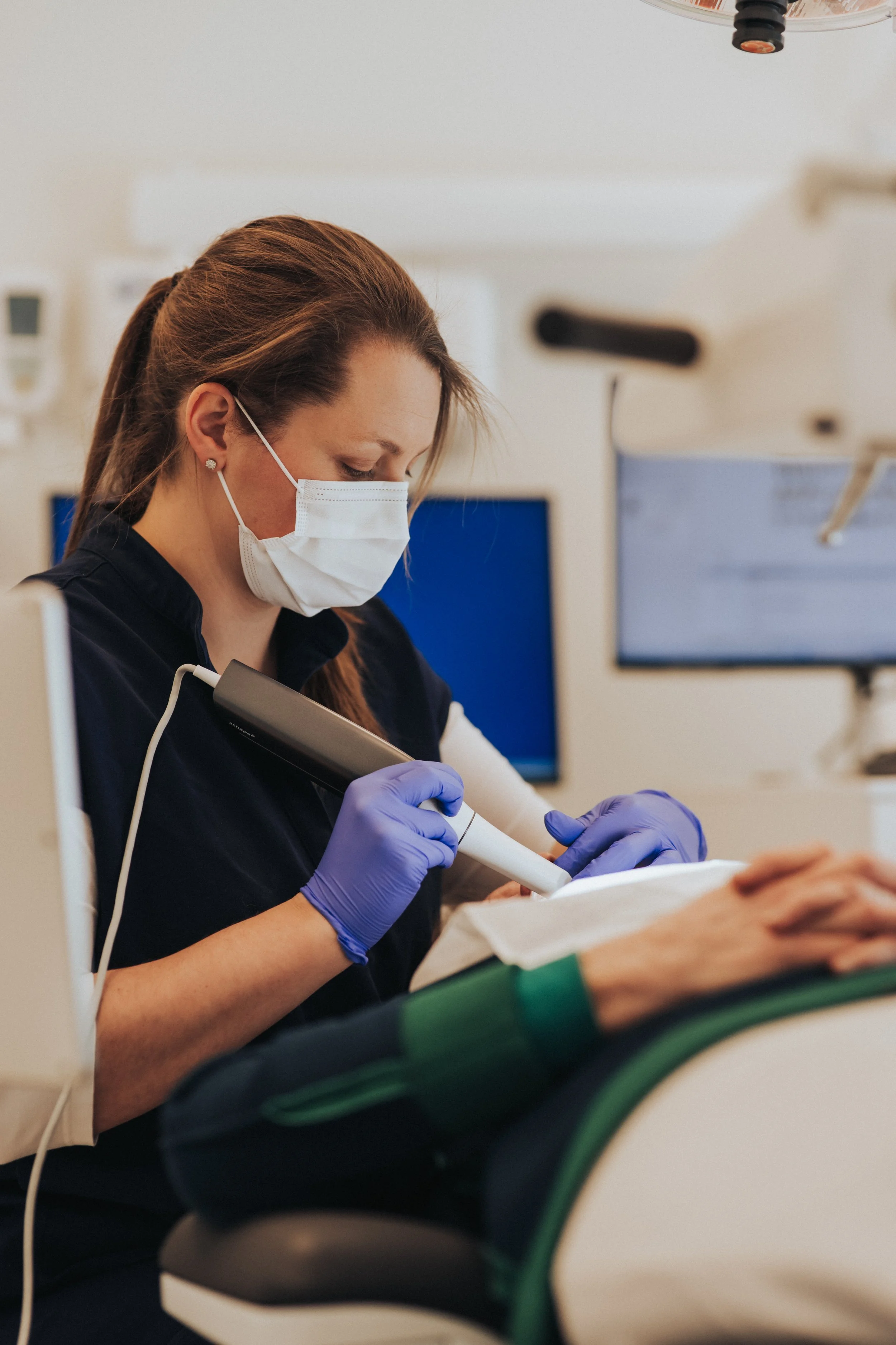 A female healthcare worker wearing a surgical mask and purple gloves uses a handheld device in a clinical setting with medical monitors in the background.