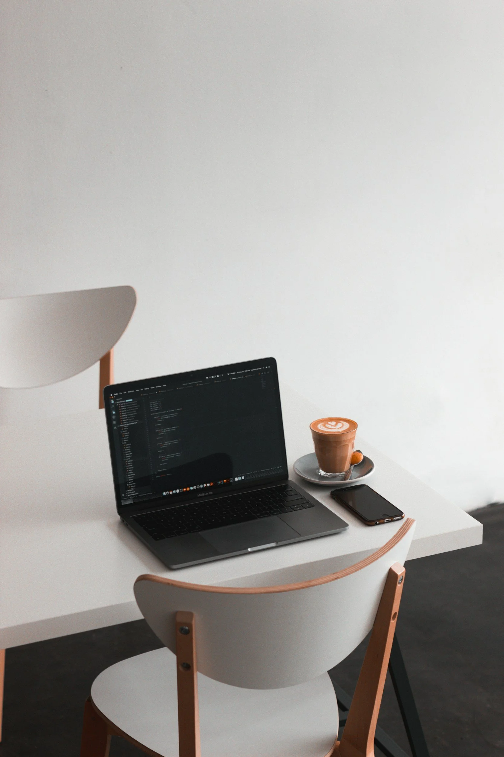 A minimalist workspace with a white desk, a chair, an open laptop showing a coding interface, a smartphone, and a coffee drink with latte art on a saucer.