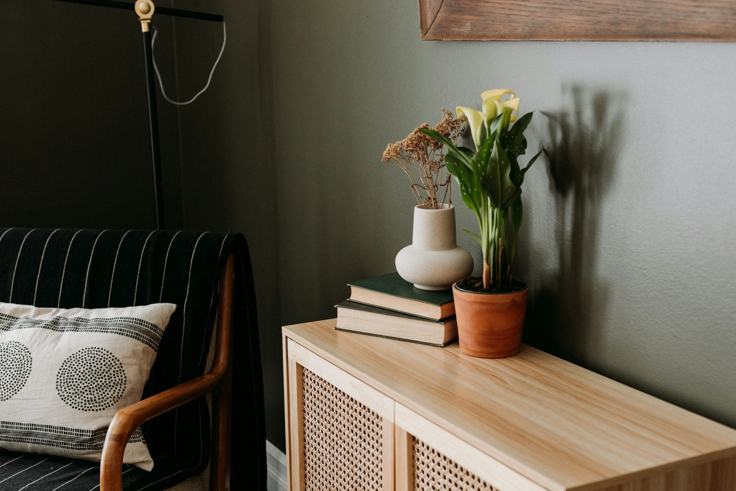 A wooden sideboard with a potted plant and a vase with dried flowers on top, next to a black-and-white striped armchair with a cushion, against a green wall.