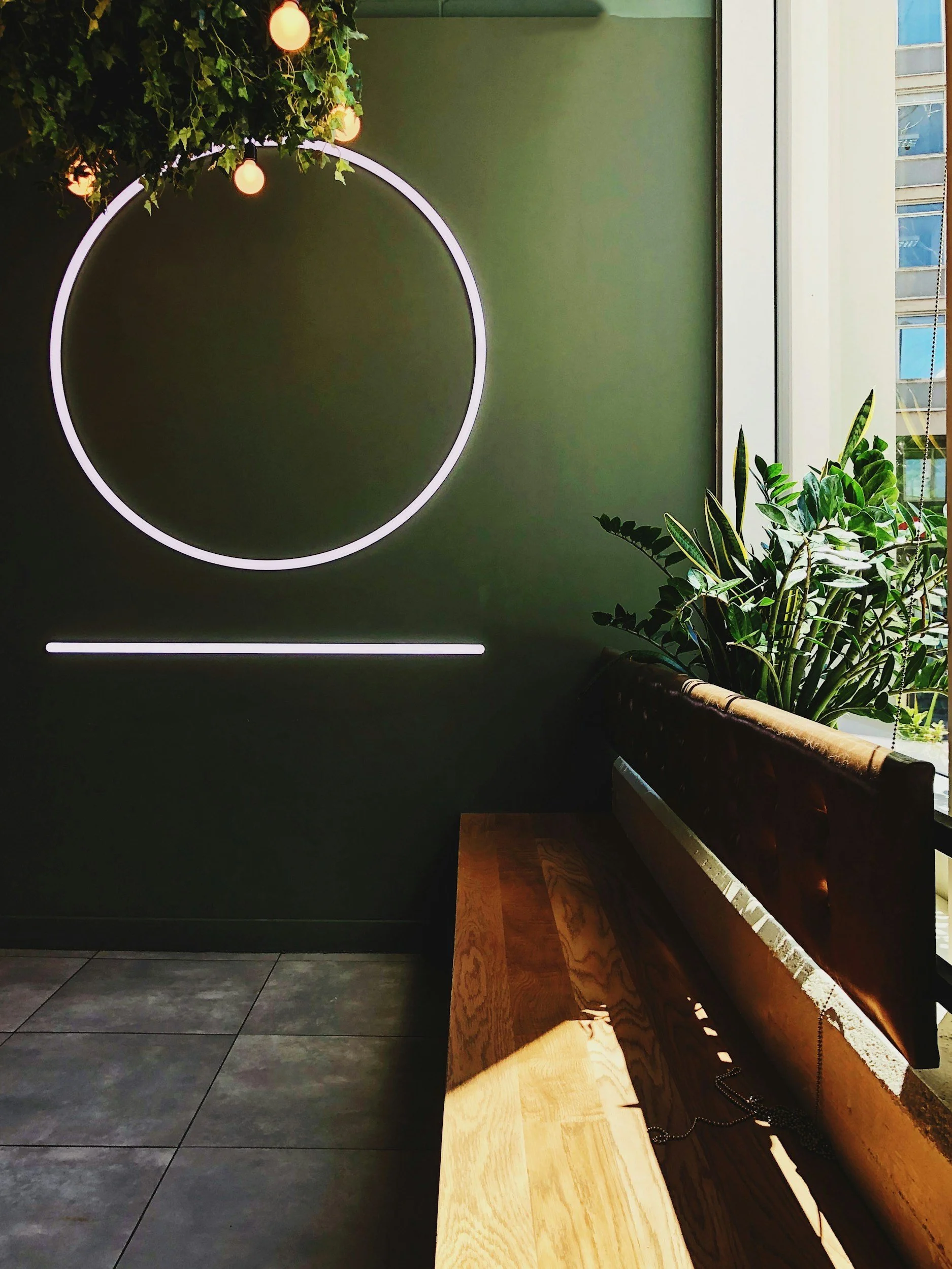 Modern interior with a neon circle and line wall art, a bench, large plant near a window, and sunlight on the tiled floor.