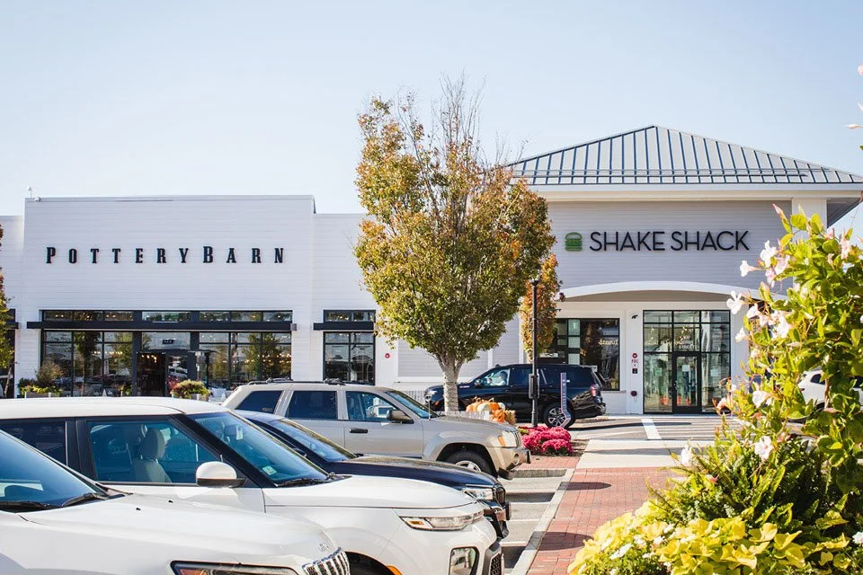 A shopping plaza with Pottery Barn and Shake Shack stores, parked cars, trees, flowers, and a clear sky in the background.