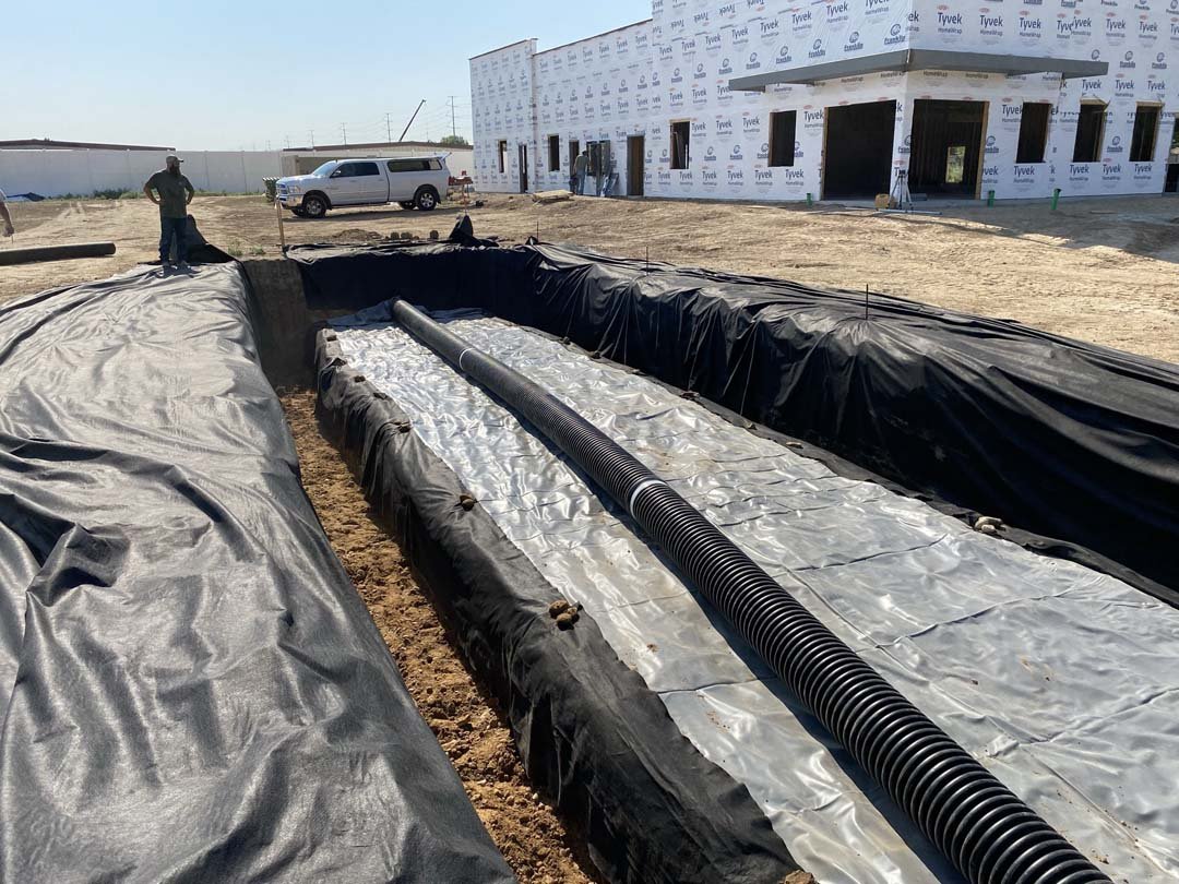 Construction site with a trench lined with plastic sheeting and drainage pipes, building under construction in the background, and a parked truck nearby.