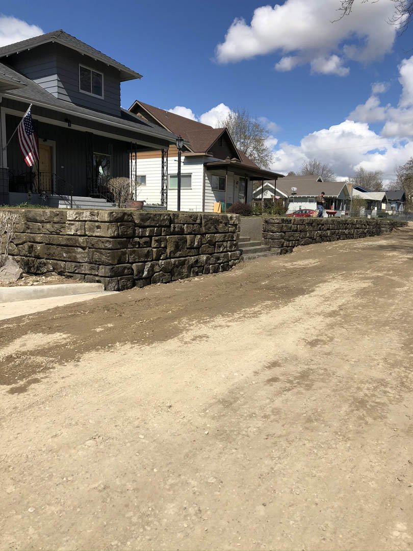 Suburban street with houses and stone retaining walls under a blue sky with clouds.