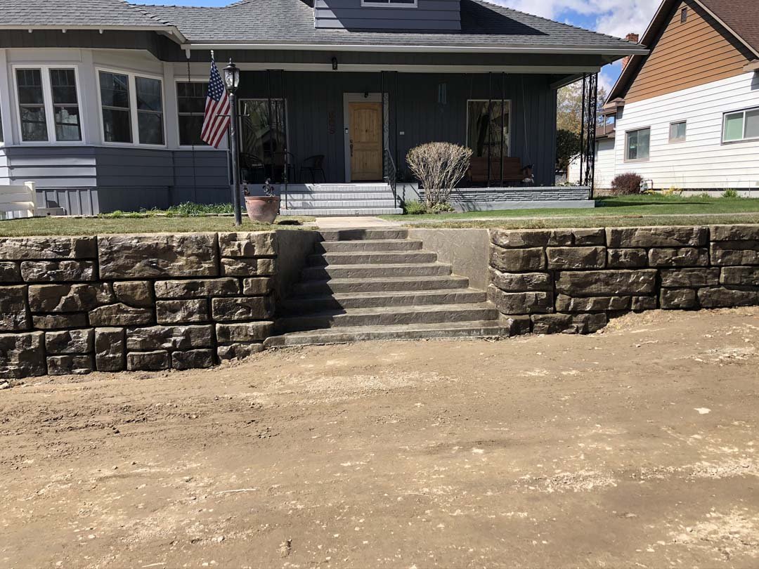 Front view of a house with gray siding, a front porch, and an American flag. Stone retaining wall with steps leading up. Partly cloudy sky.