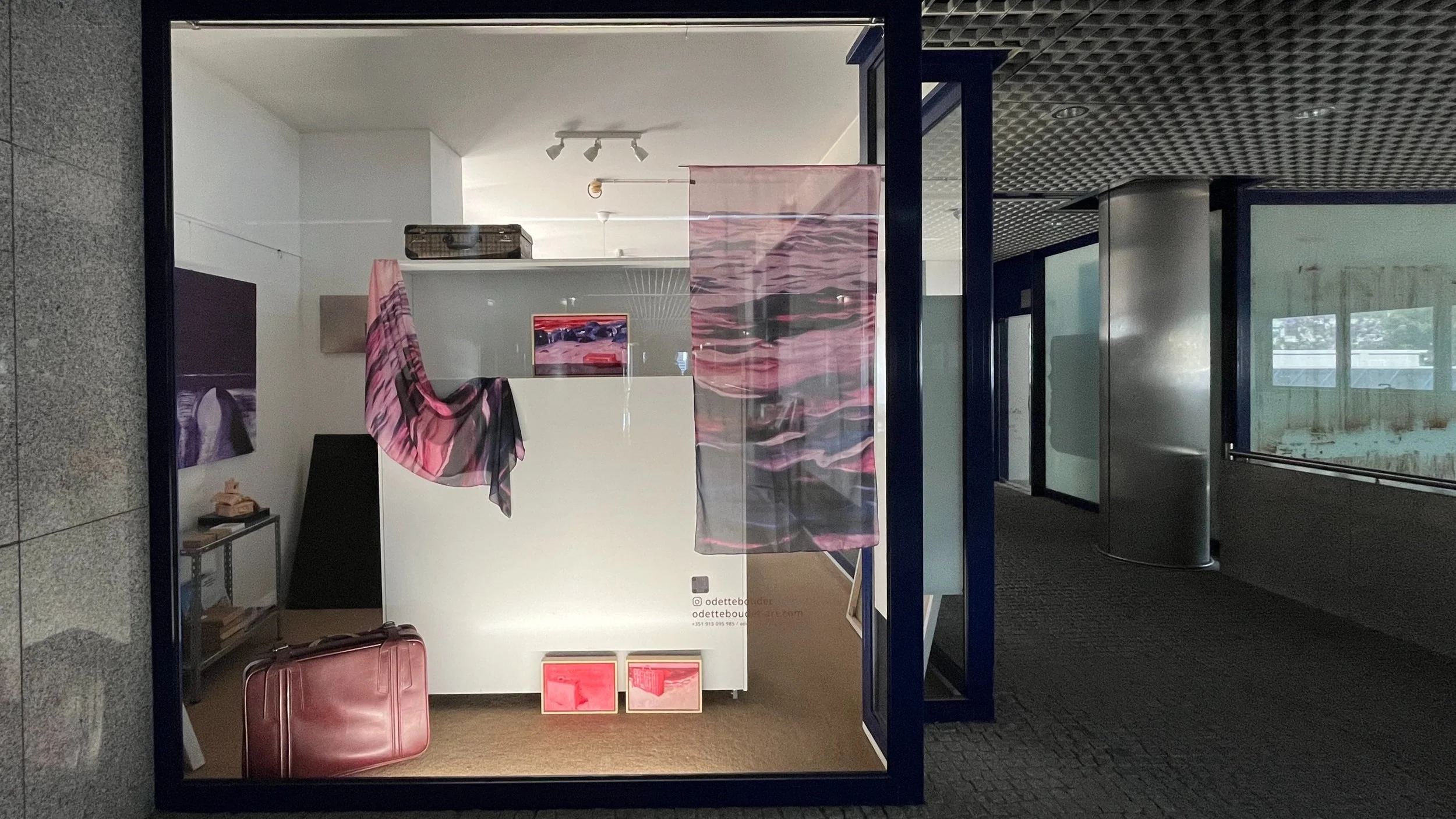 Sala de exposição com roupas coloridas de tecido suspensas, uma mala de viagem rosa escura no chão, uma mesa de metal com objetos e duas obras de arte com cores semelhantes ao tecido, mostrandose do lado de fora de uma loja ou galeria.