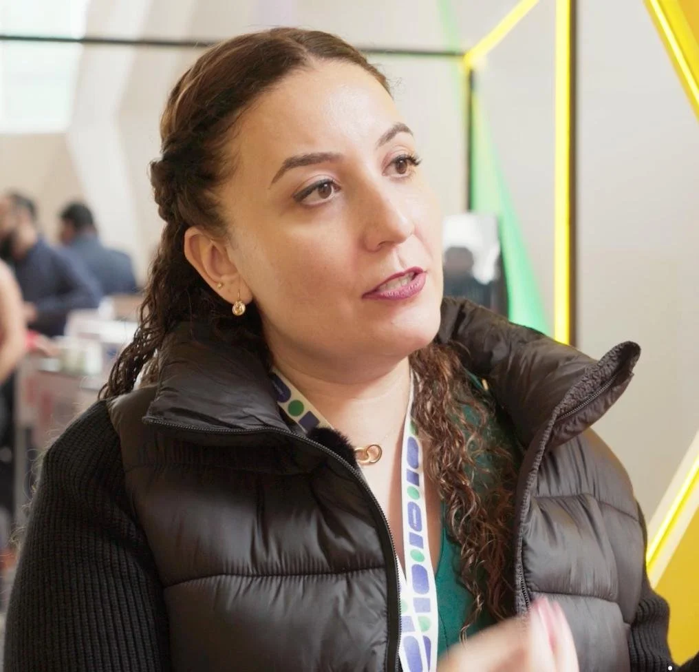 Mujer con cabello rizado y piel clara en una conferencia o evento, con fondo de otras personas y decoraciones modernas.