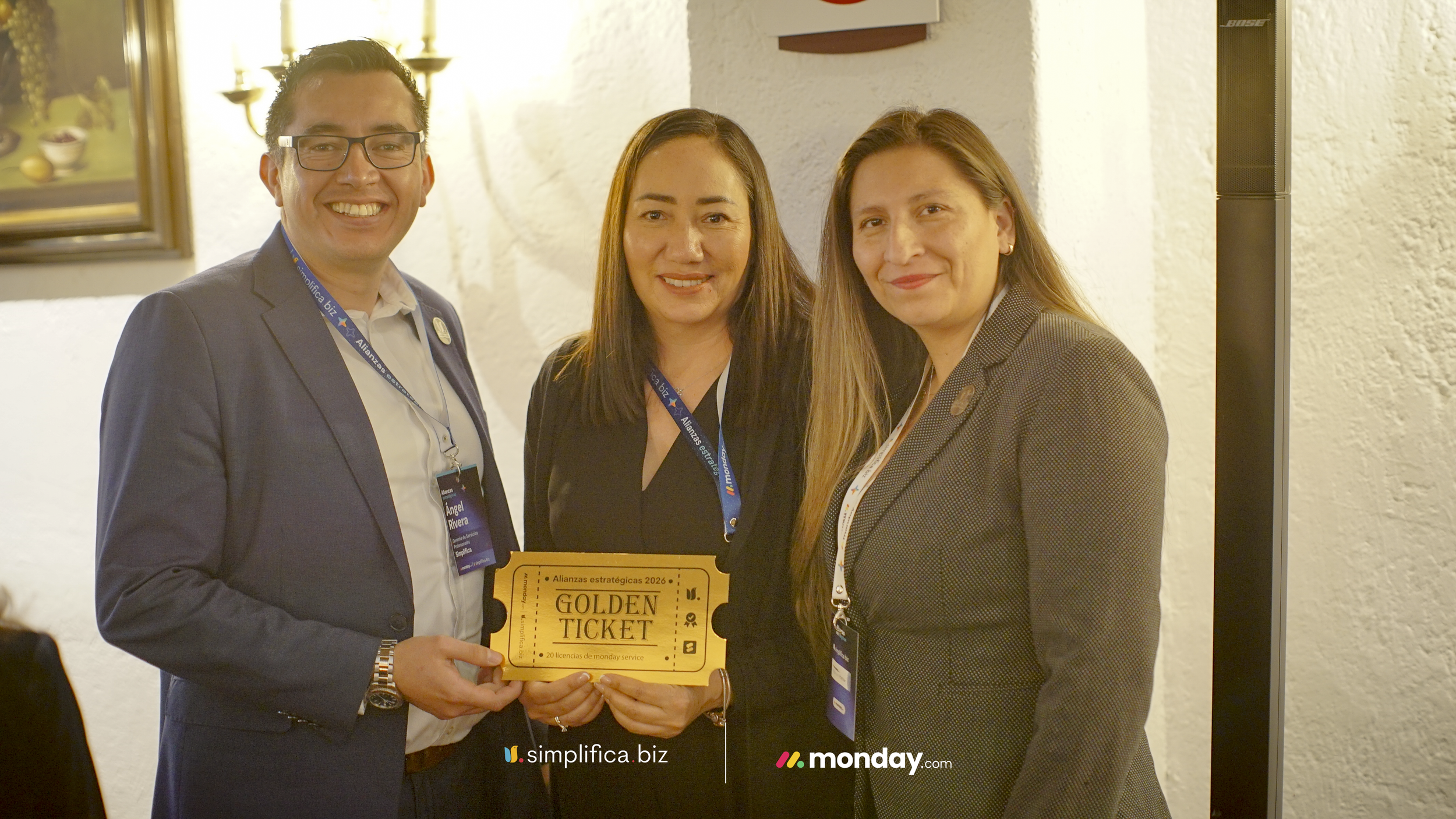 Tres personas de pie en un evento, la persona del centro sostiene un boleto de premio dorado que dice "Golden Ticket" y tiene credenciales colgadas en cuello, todos son sonriendo y viste traje formal.