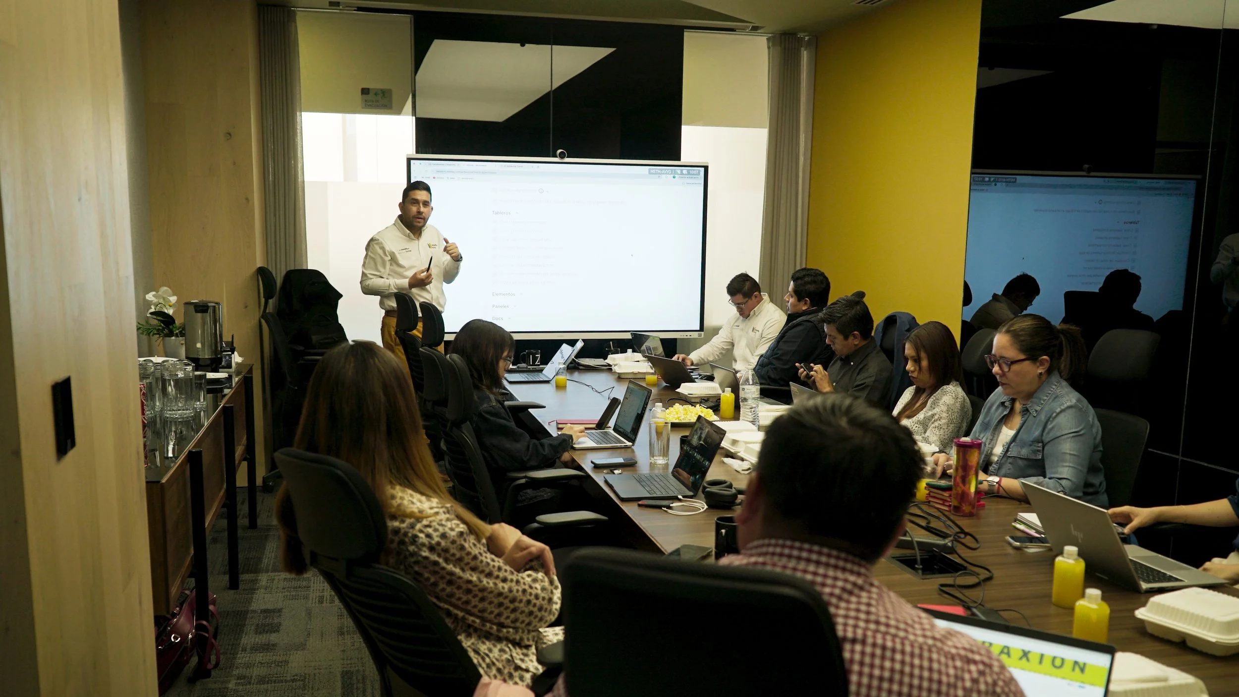 Reunión de negocios con varias personas en una sala de conferencias, algunas usando computadoras portátiles, escuchando a un presentador delante de una pantalla grande.
