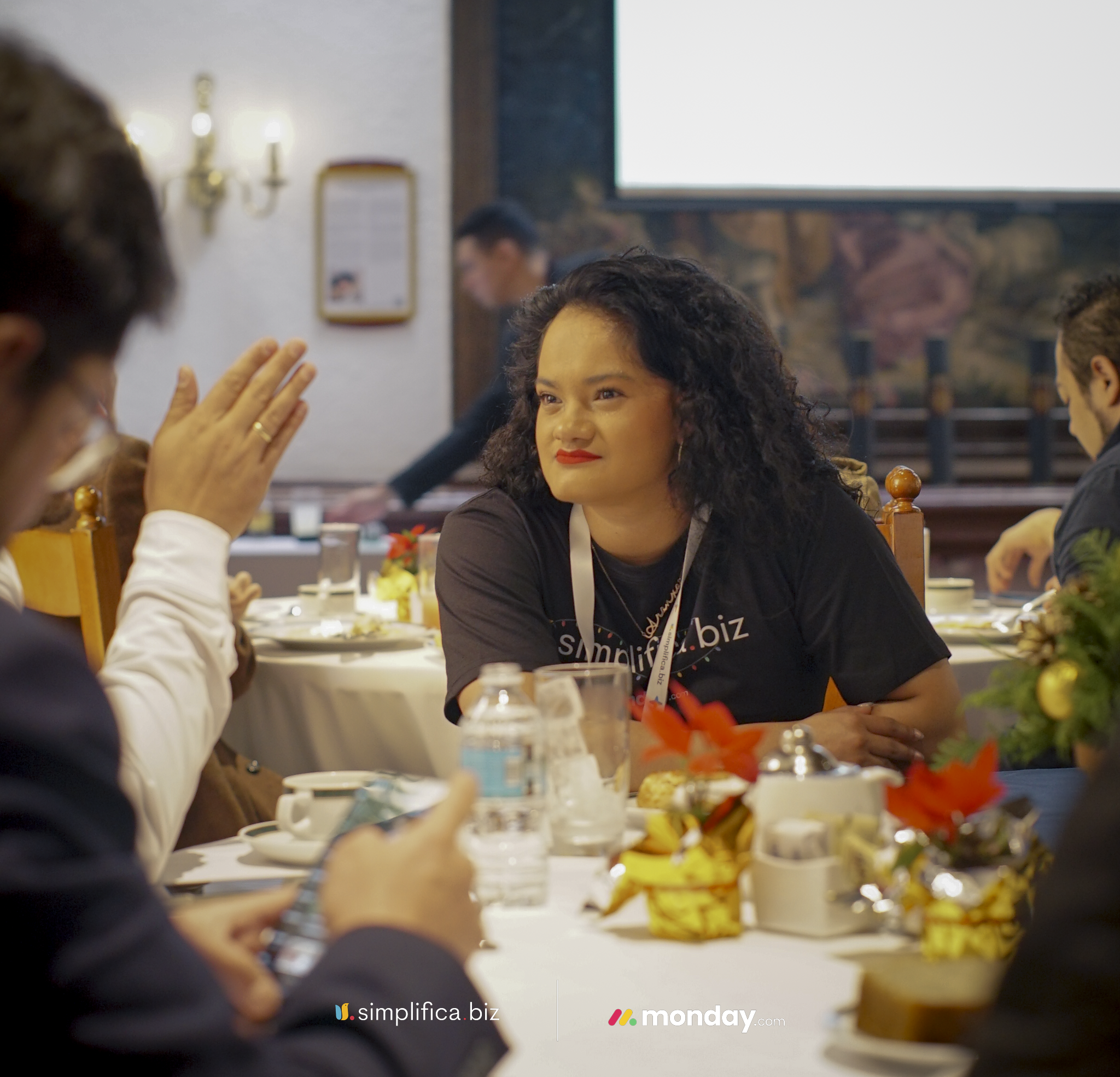Grupo de personas en una comida o reunión social, sonriendo y conversando en un ambiente decorado con flores y objetos como botellas y platos de comida en una mesa.