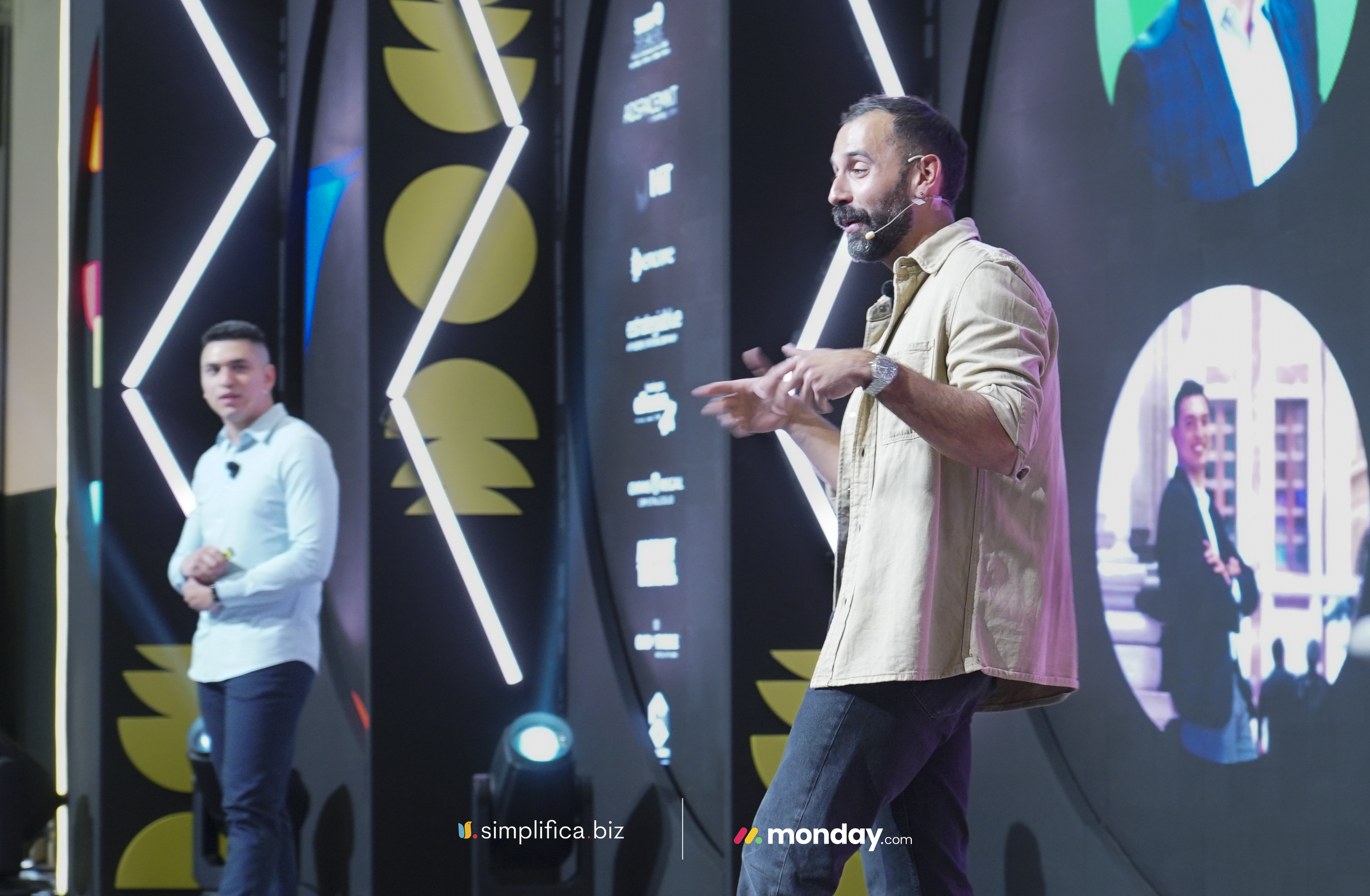 Hombre de pie hablando frente a una pantalla grande con varias personas en el fondo en un ambiente moderno y tecnológico.
