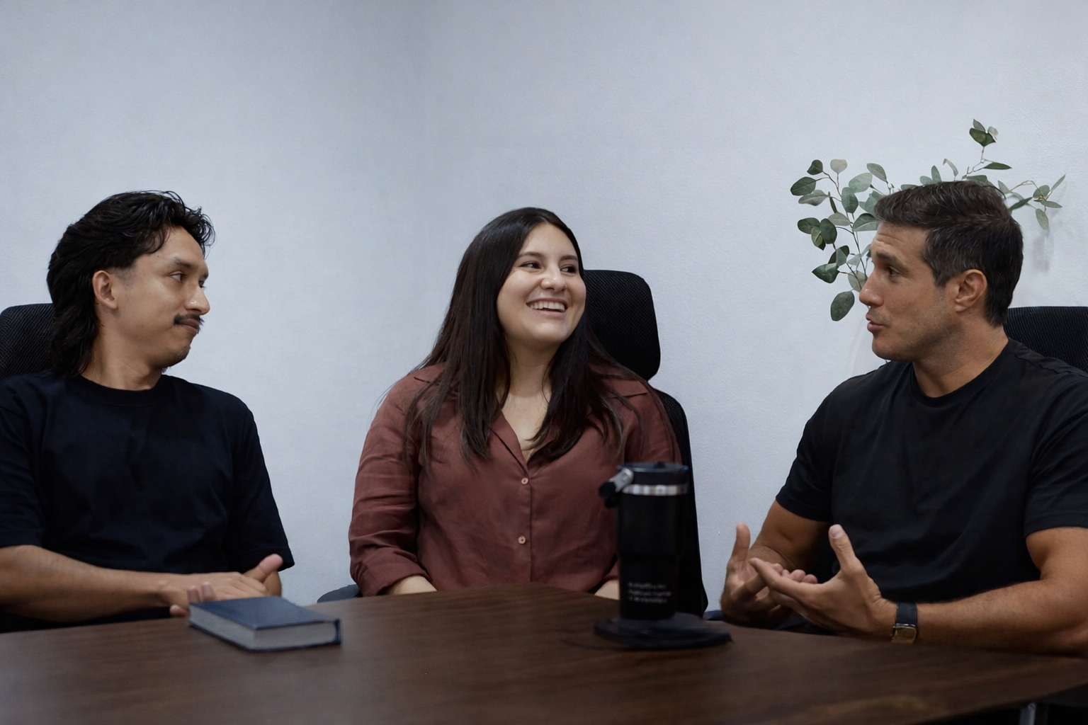 Tres personas en una reunión de trabajo, dos hombres y una mujer, sentados alrededor de una mesa, conversando y sonriendo.