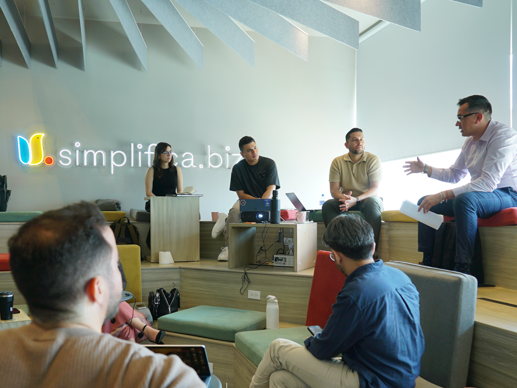 Reunión en una oficina con varias personas, un hombre en el extremo derecho en una silla de color naranja está hablando, mientras los demás escuchan. Hay un logotipo en la pared que dice 'simplifica.biz' y una pantalla con un proyector en una mesa.