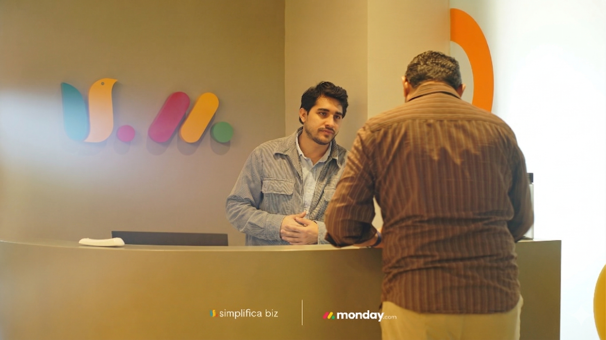 Dos hombres en una oficina conversando en un mostrador. Detrás, un logotipo colorido y la marca 'simplifica biz' y 'monday.com' en la parte inferior de la imagen.