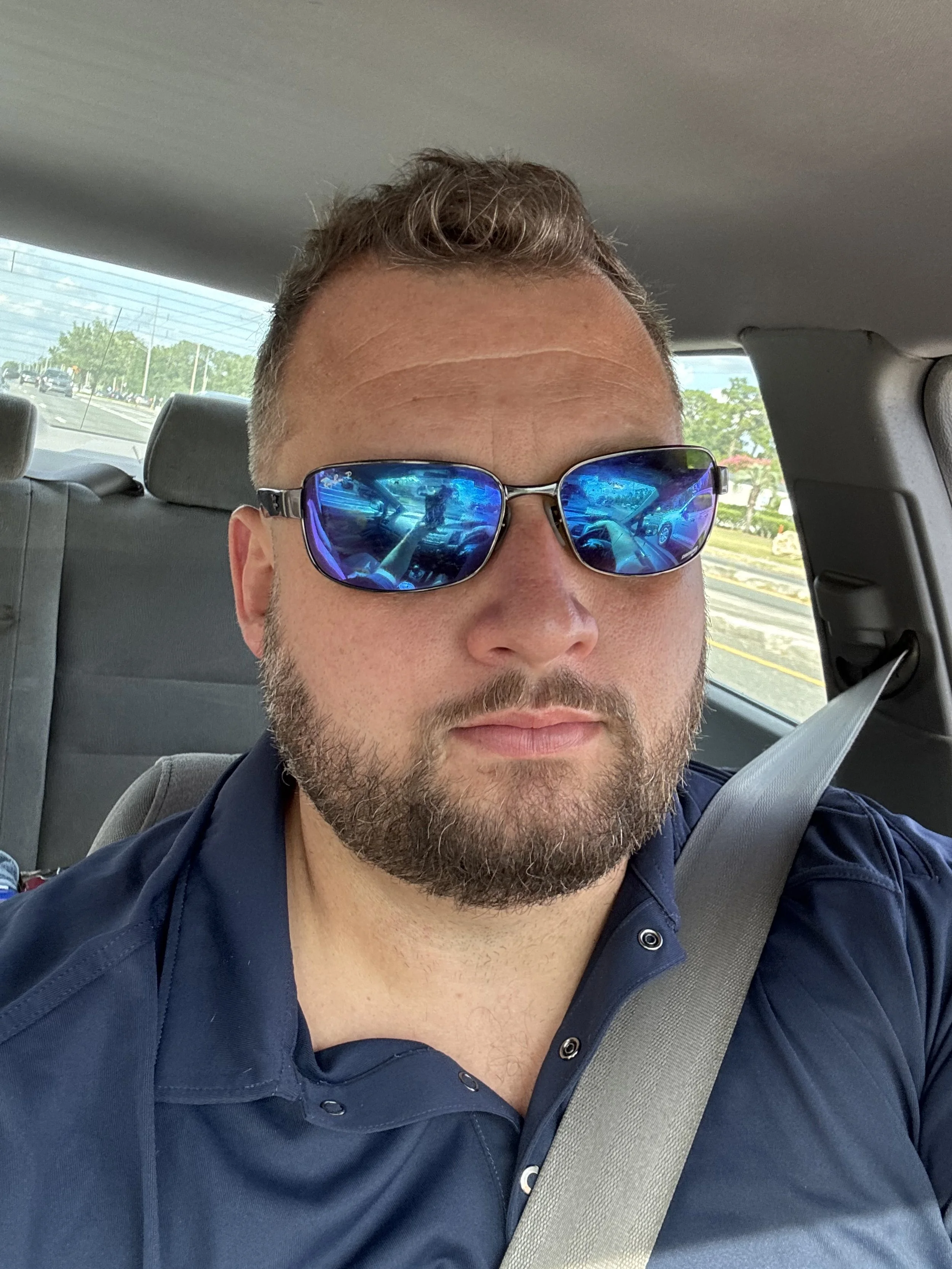 A man with a beard wearing blue reflective sunglasses and a navy blue shirt, sitting in a car with a seatbelt on.