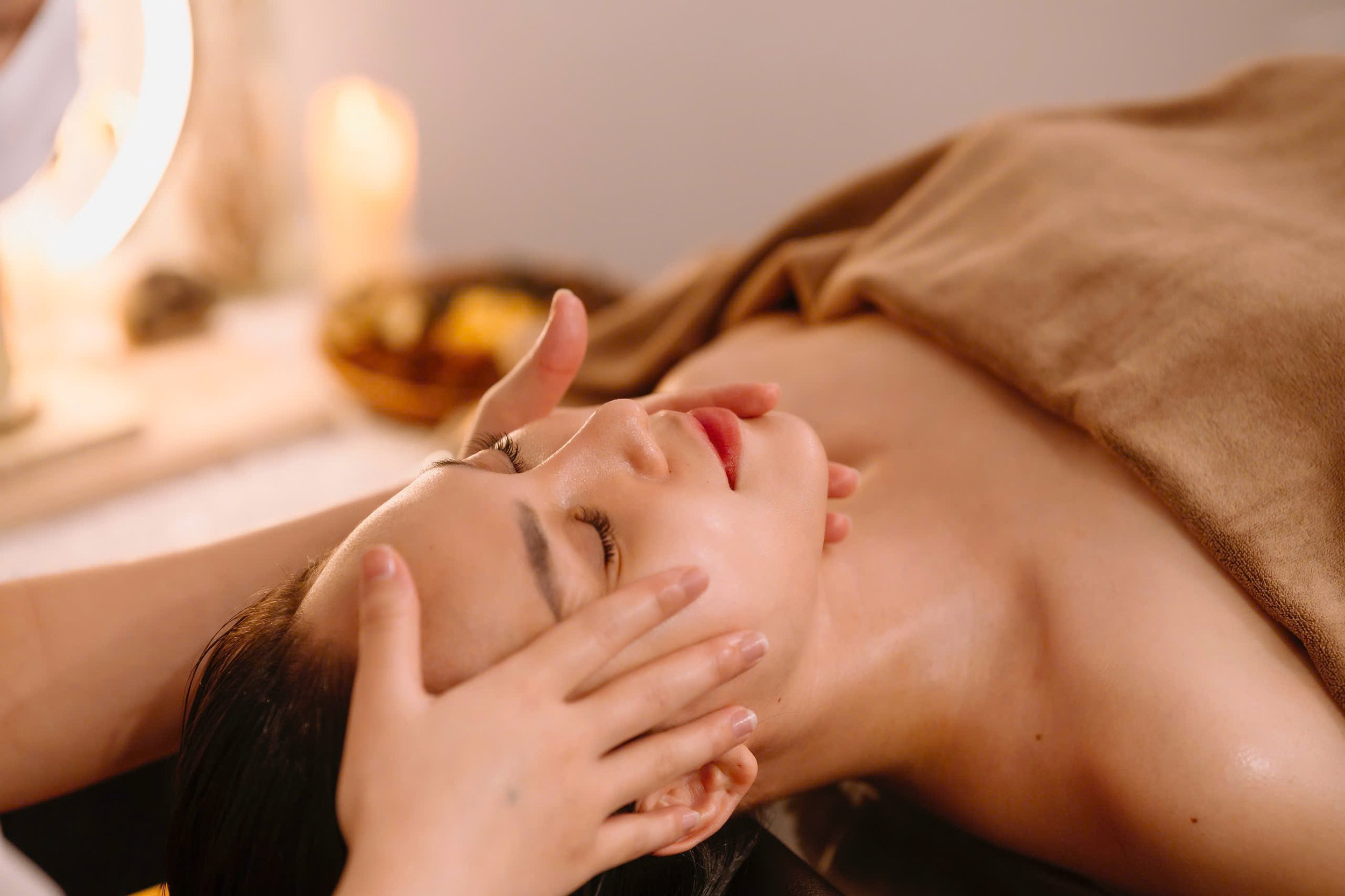 Spa treatment with woman receiving facial massage, covered with a brown towel.