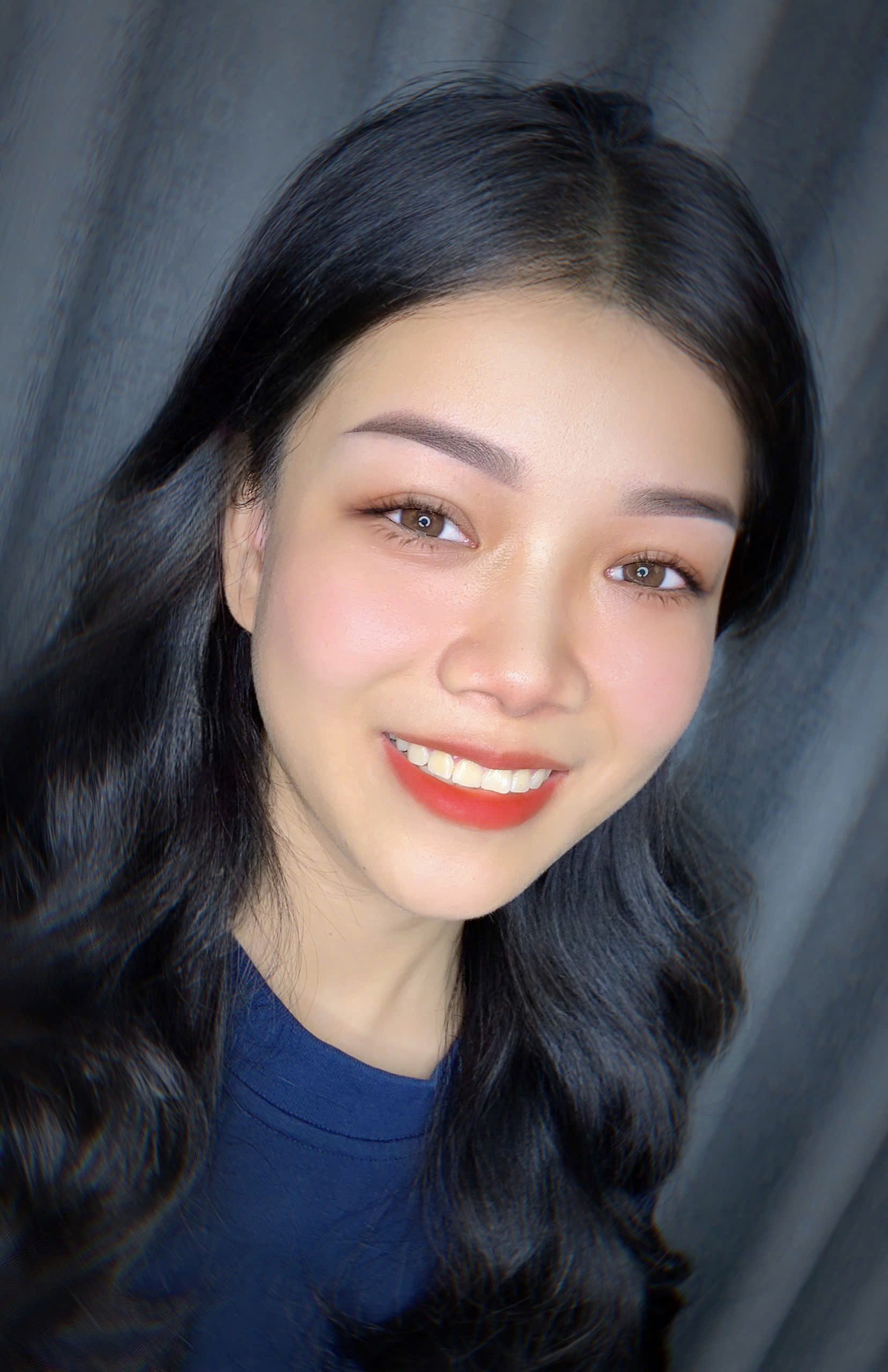 A woman with long black hair smiling, wearing a blue shirt, against a gray background.