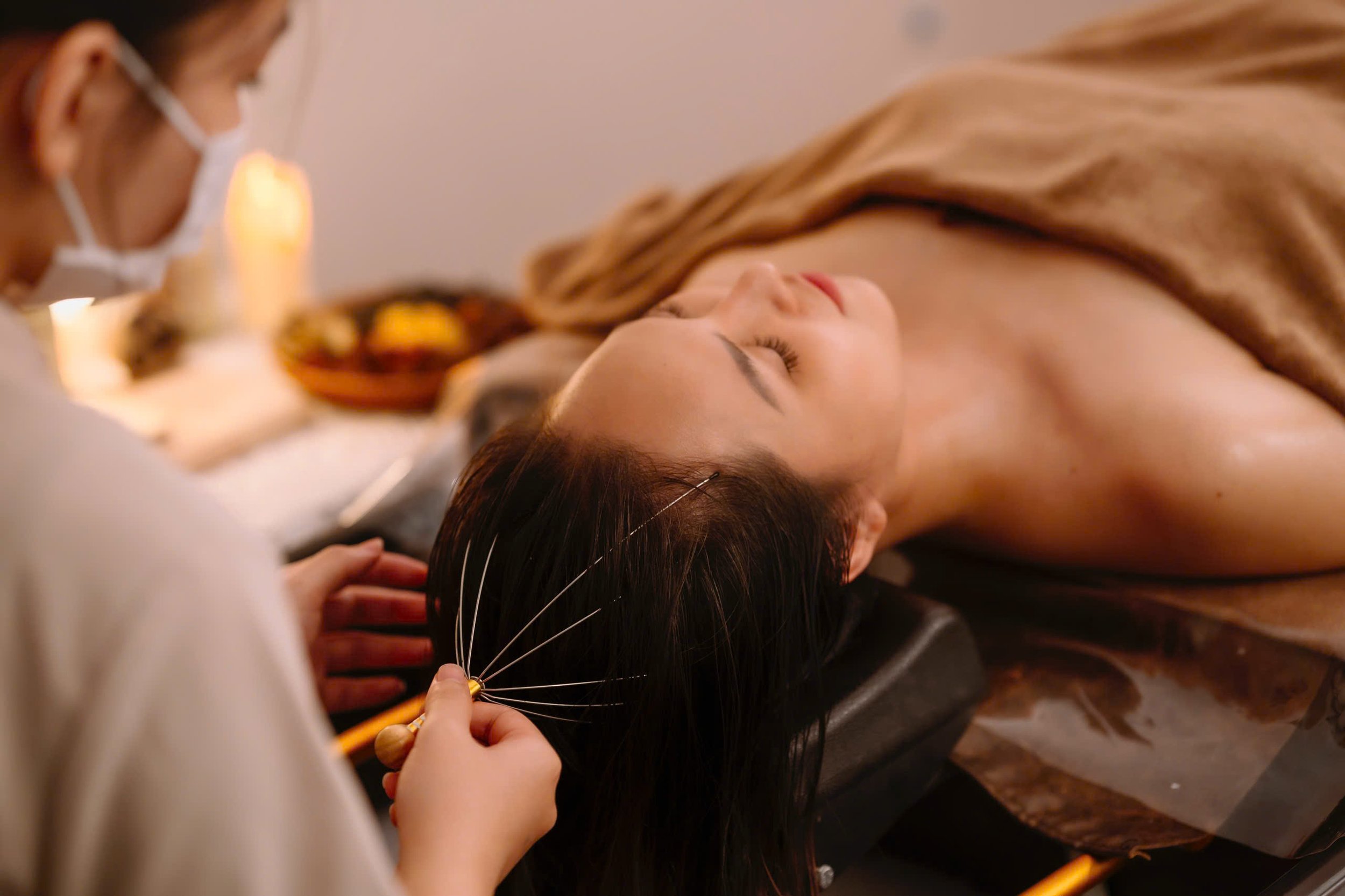 Person receiving a head massage with a scalp massager tool in a spa setting, relaxing under a towel.