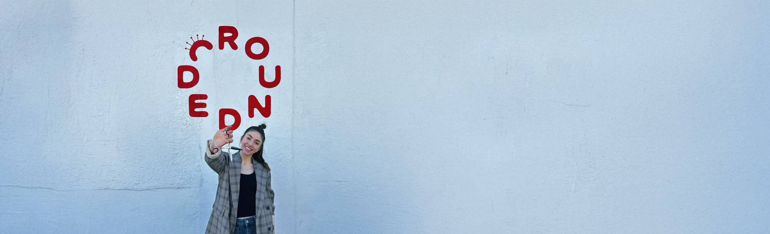A woman holding a pair of scissors, standing in front of a light blue wall with red letters spelling 'ROUNDED'.