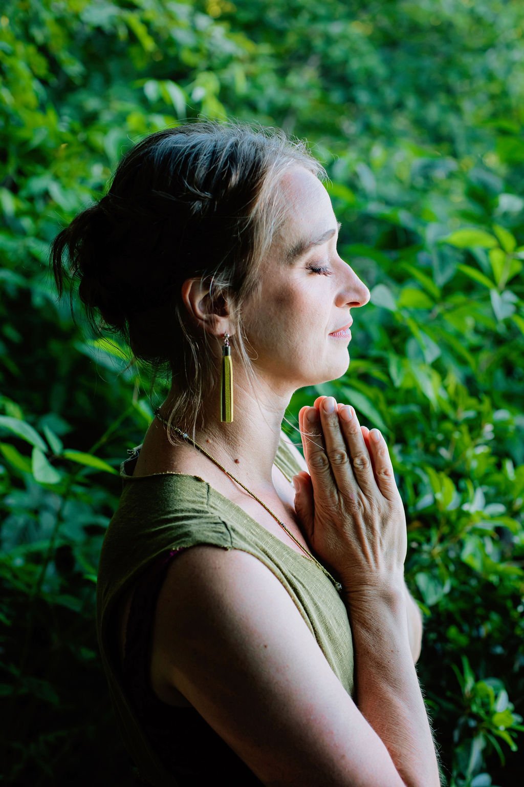 A woman with closed eyes and clasped hands in prayer, standing against a green leafy background.
