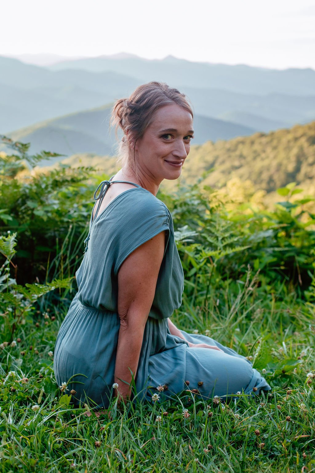 A woman in a blue dress sitting on green grass with a backdrop of rolling green mountains under a cloudy sky.
