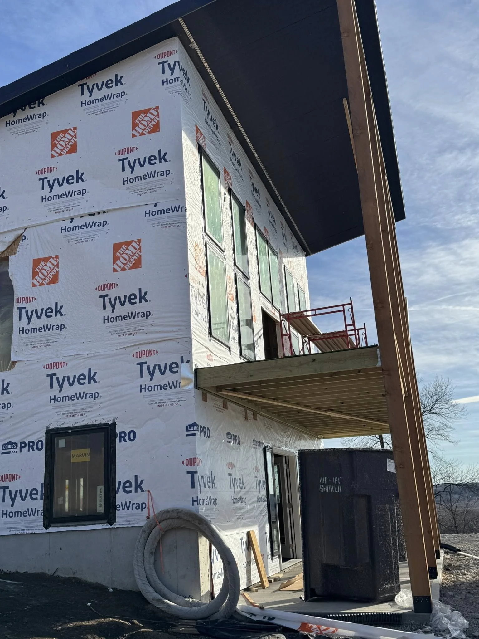 Building under construction wrapped in Tyvek HomeWrap with multiple windows installed, a small overhang, and construction materials and equipment visible in the foreground.