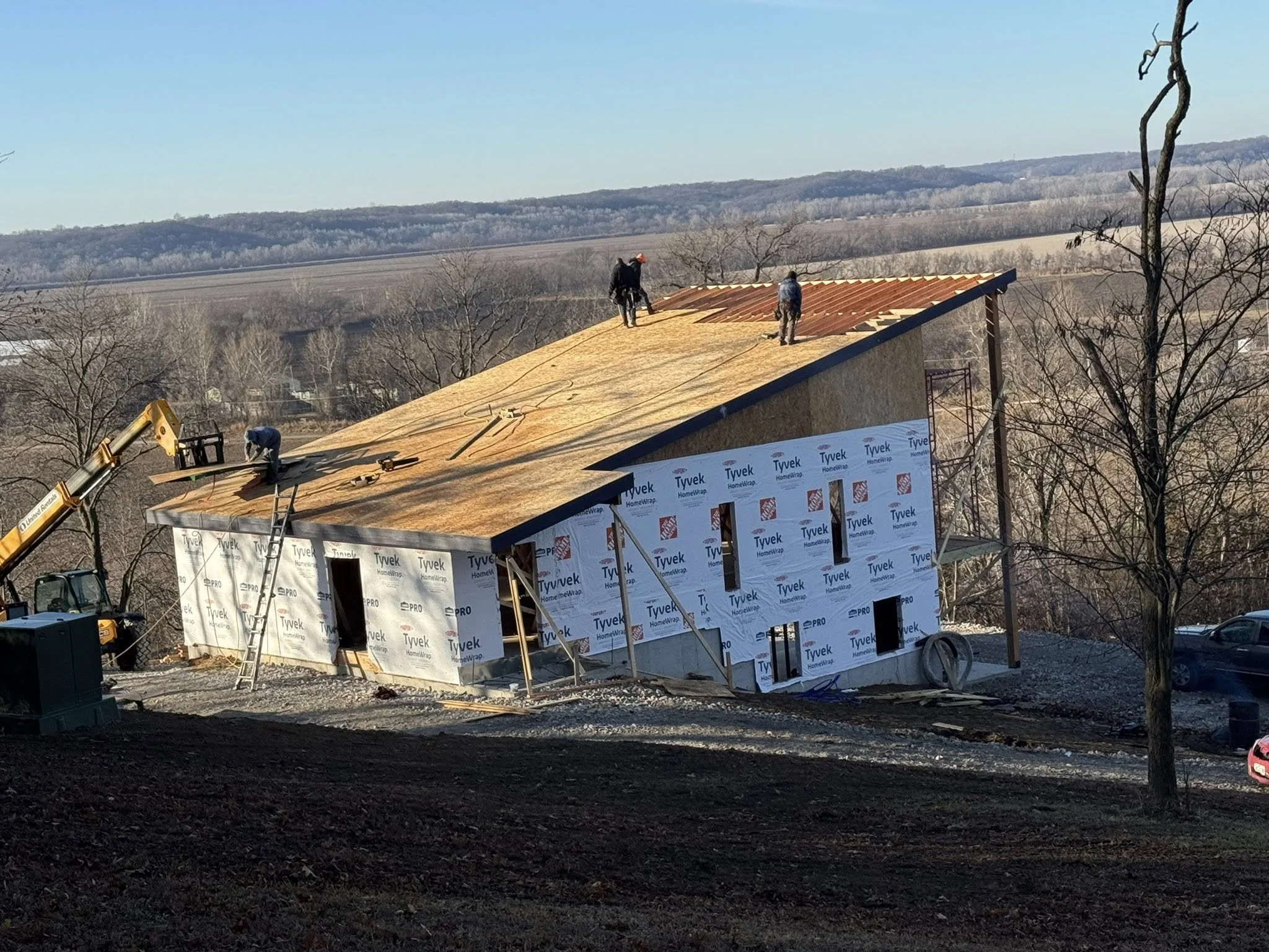Construction workers installing a metal roof on a house, with some workers working on the roof and others on the ground, surrounded by trees and distant hills.