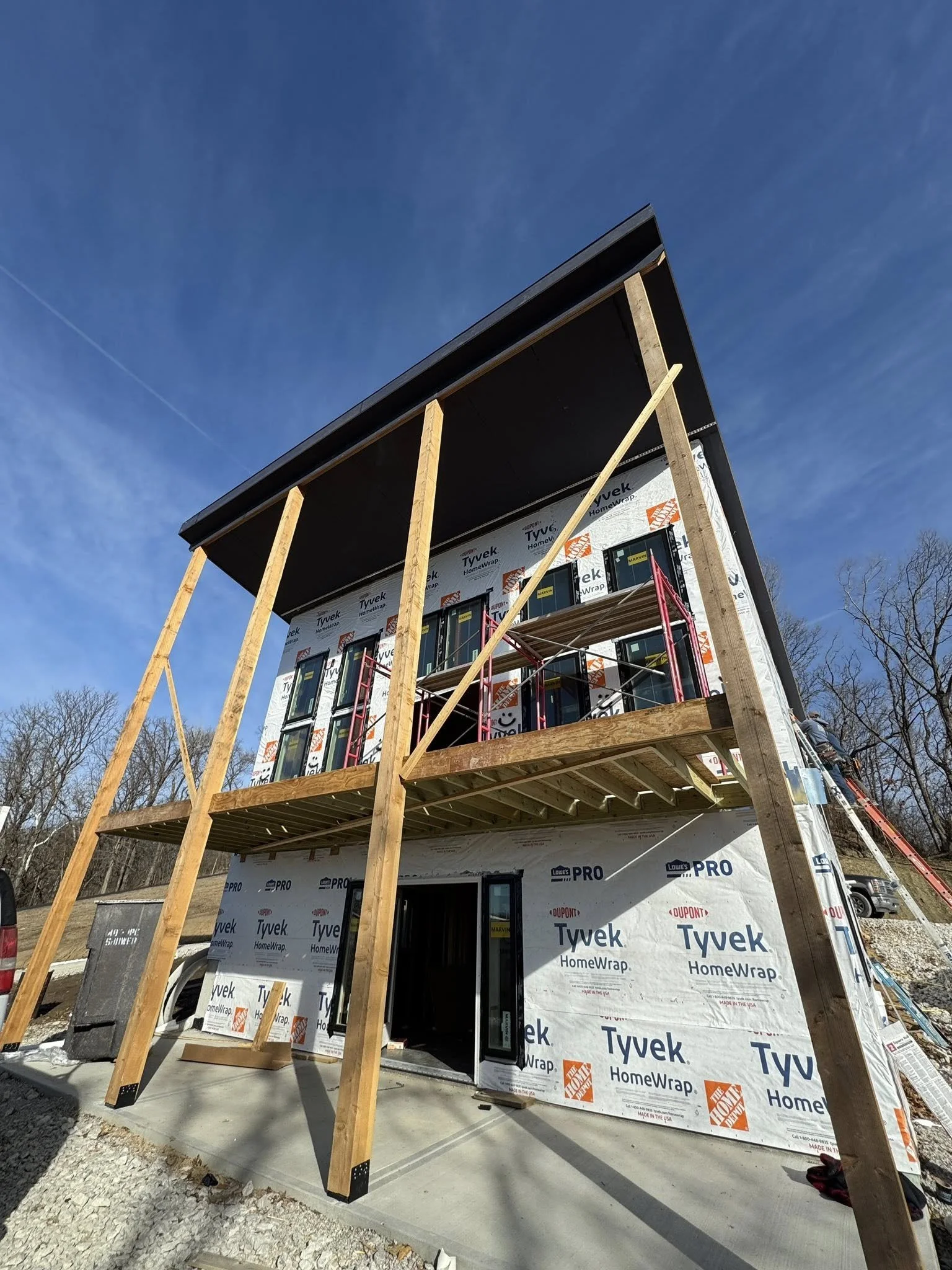 Under-construction two-story house with exposed wooden framing, wrapped in Tyvek HomeWrap, and scaffolding on the second floor. The house has black roofing, and construction workers are present. The sky is clear and blue.