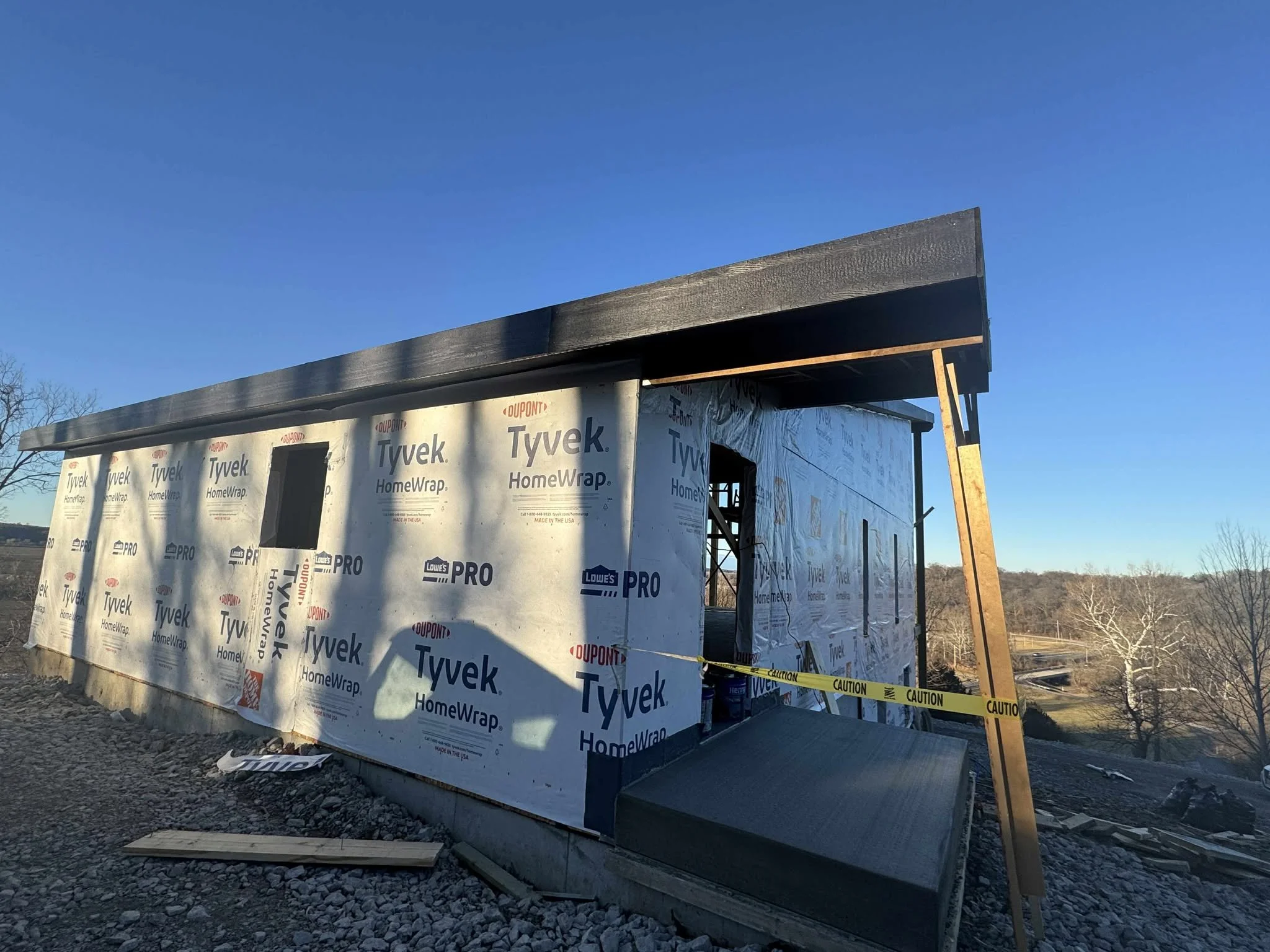 Construction site with a small building under construction, wrapped in Tyvek HomeWrap, with a raised ramp and caution tape in front, and a blue clear sky in the background.