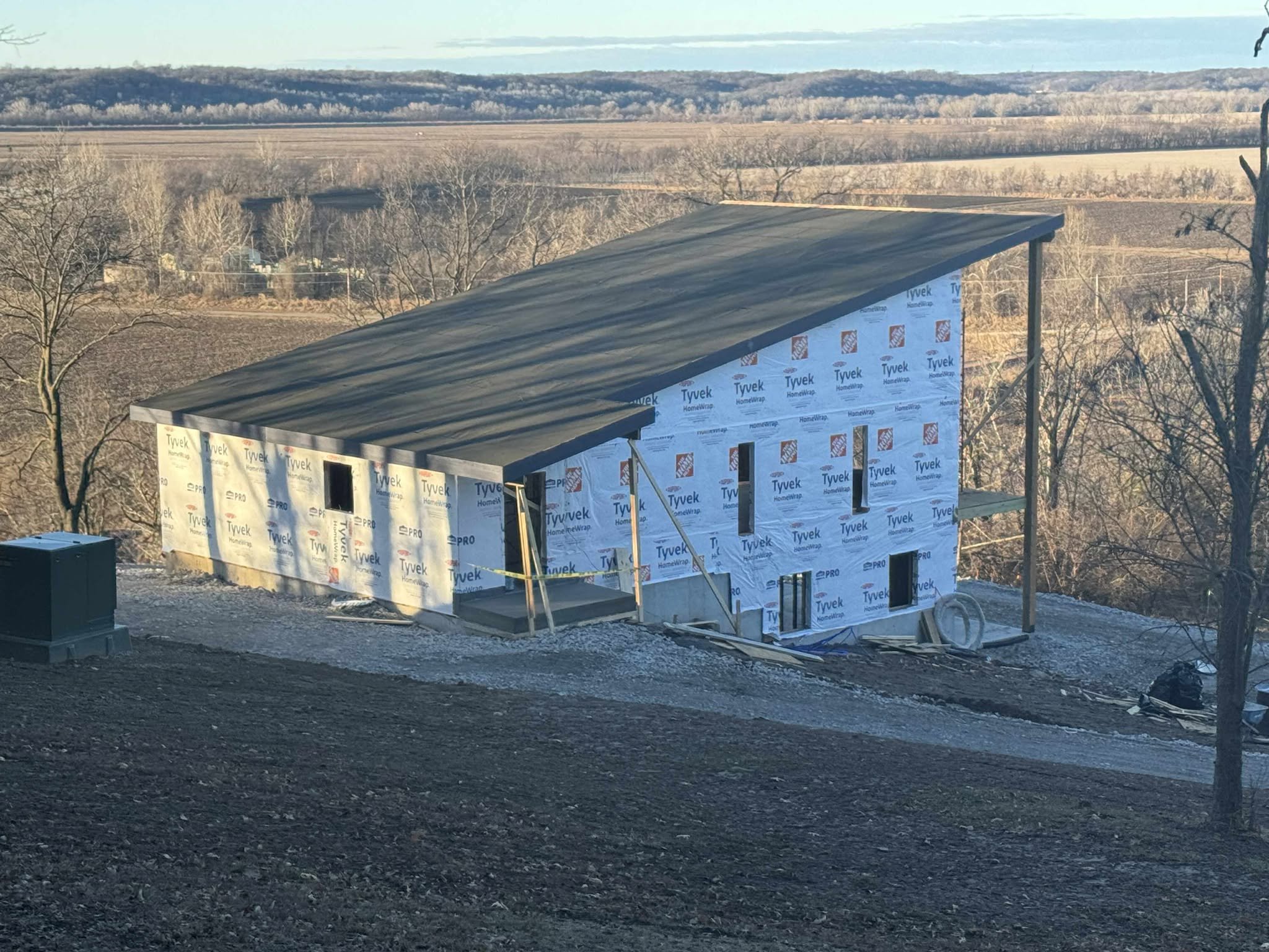 Under construction house with Tyvek house wrap, windows not yet installed, and a sloped roof, set in a rural landscape with trees and open fields in the background.