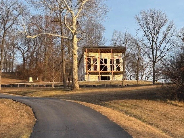 A partially constructed two-story wooden house on a hilltop with leafless trees around, a paved winding road in the foreground, and a clear sky.