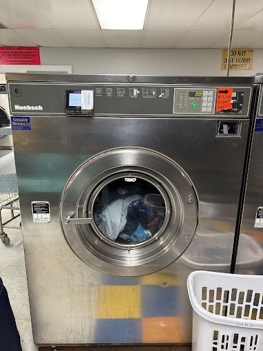 Commercial washing machine with clothes inside, located in a laundromat, next to a white laundry basket.