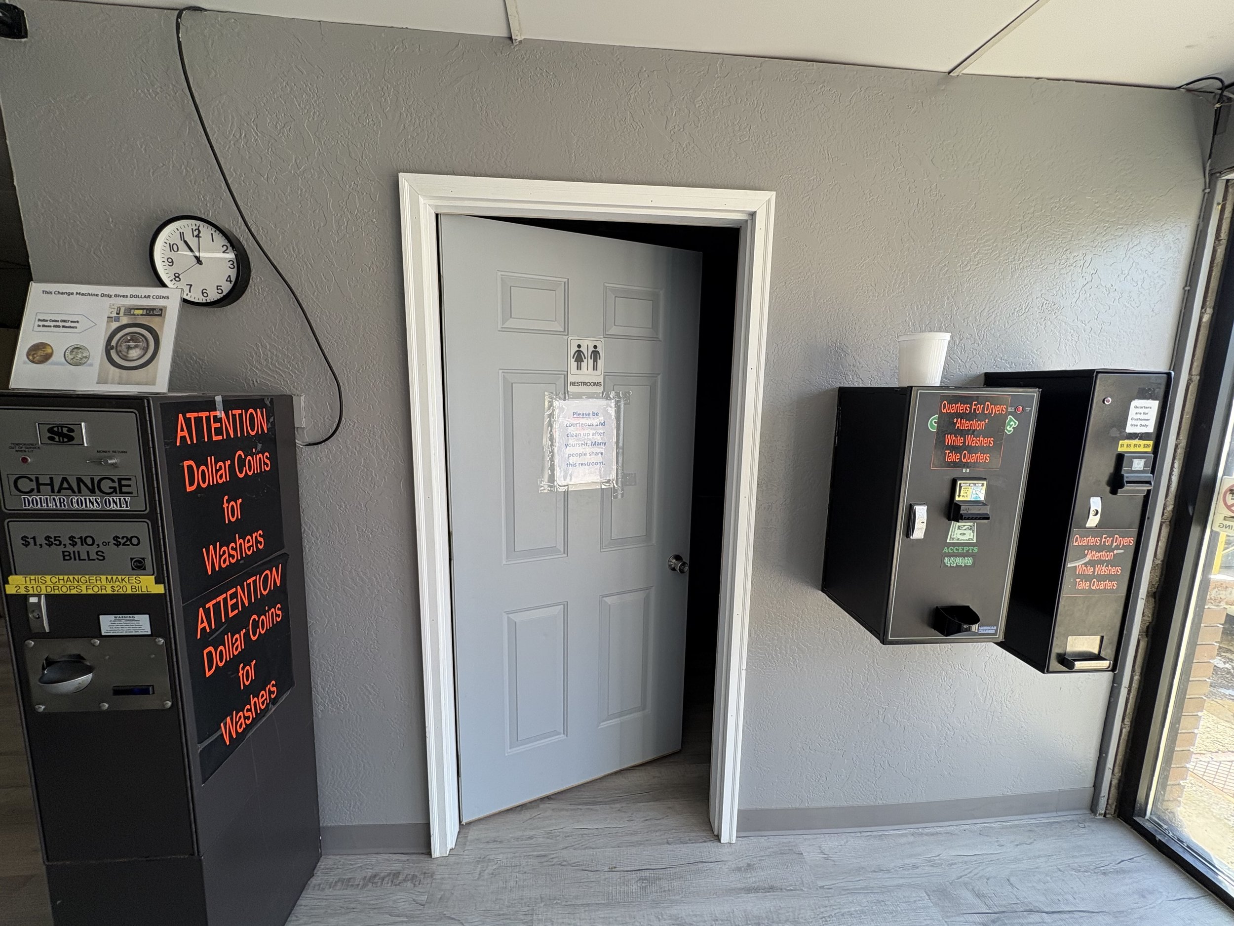 Interior of a laundromat with a coin change machine, a restroom door, and signage stating use of dollar coins for washers and quarters for dryers. A clock is mounted on the wall, and a transparent cup is placed on a ledge.